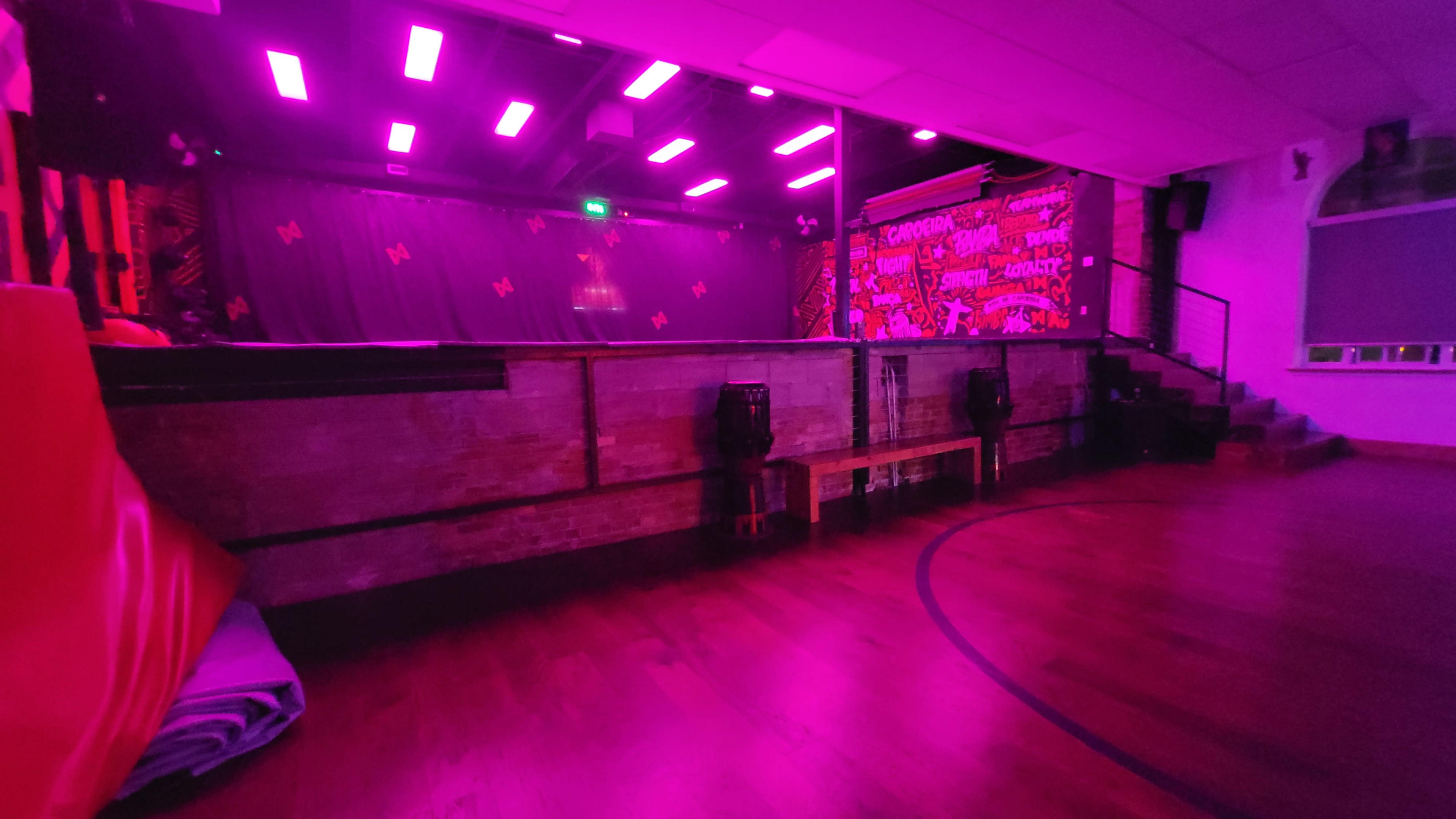 The image shows a dimly lit indoor space with purple lighting, featuring a stage with a decorative backdrop and empty seating.