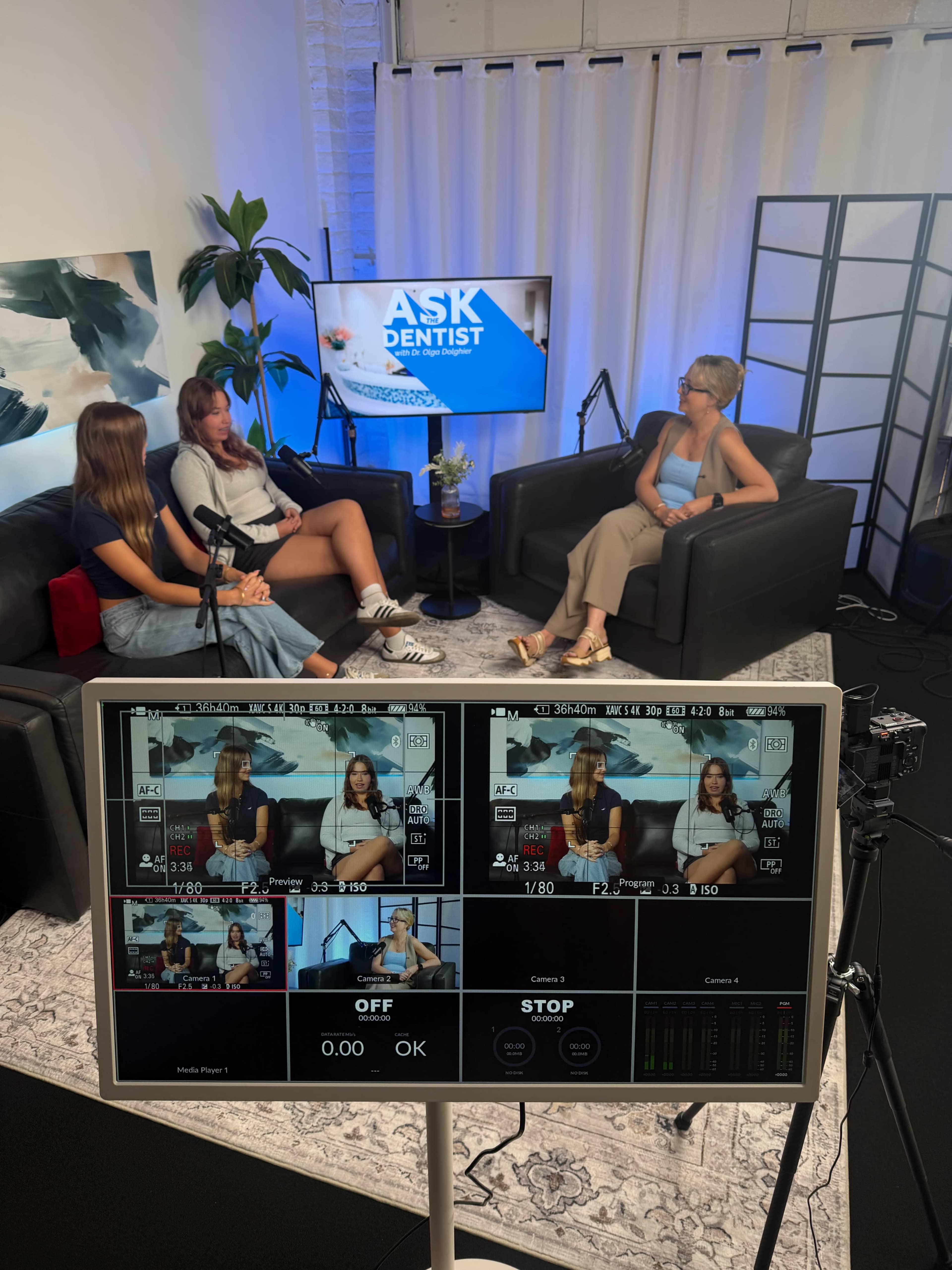 The image shows a recording studio setting where three women are seated on black couches discussing a topic, with a monitor displaying the scene and text reading "Ask Dentist."