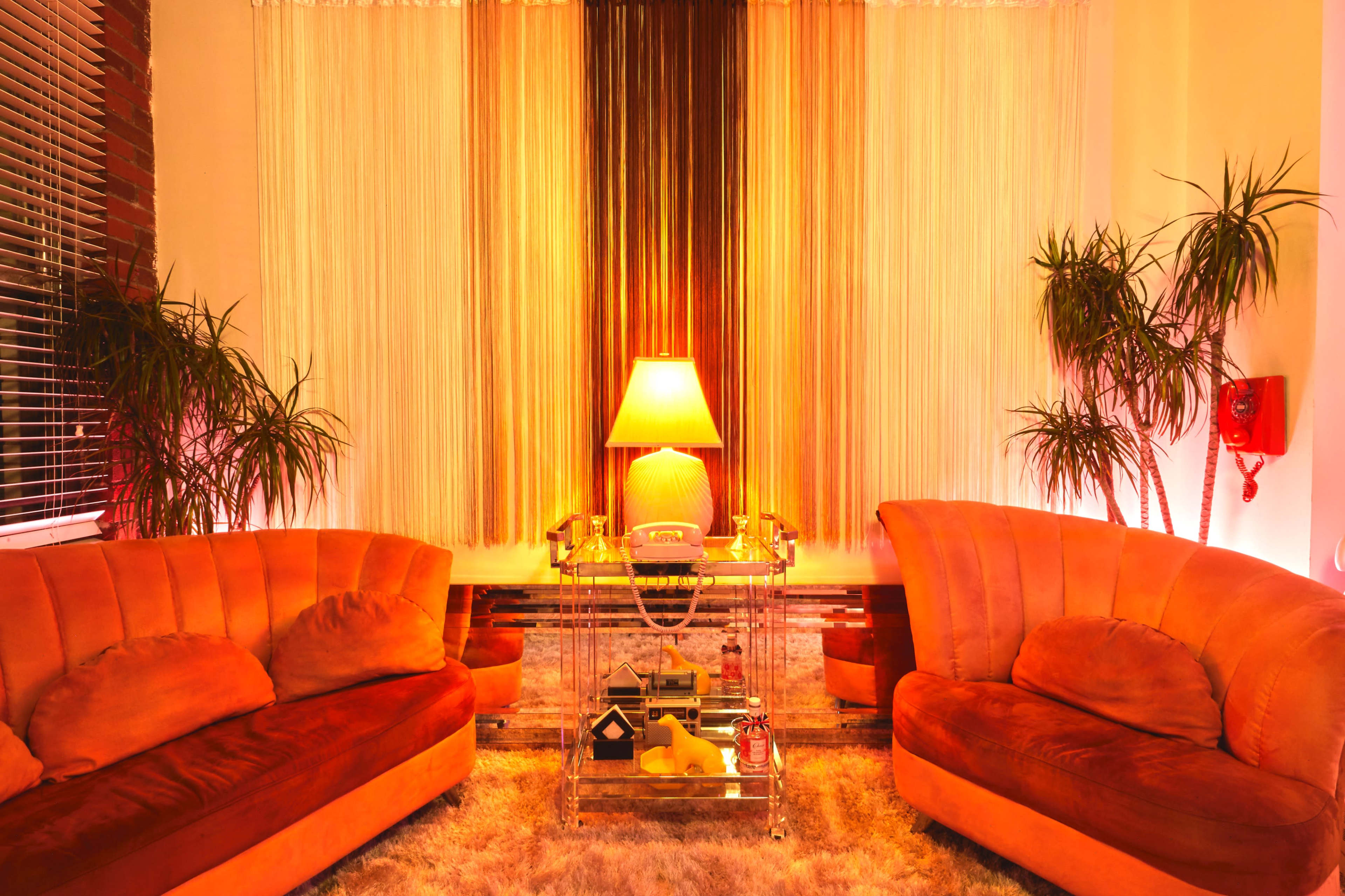 A cozy living room with two orange sofas, a glass coffee table, a lamp, and potted plants, all illuminated by warm lighting.