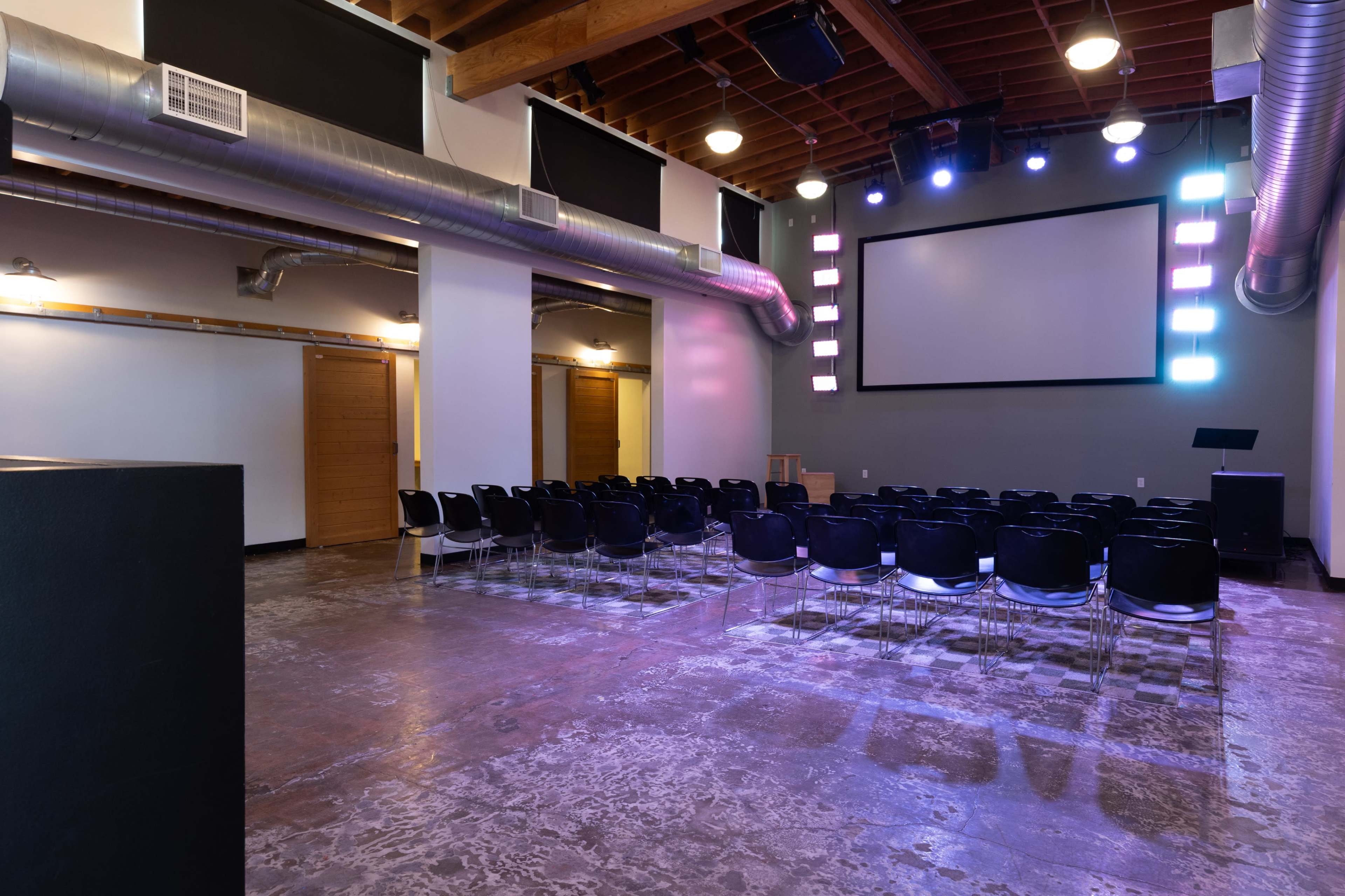 The image shows an empty, modern auditorium with rows of black chairs facing a large screen, illuminated by colored lights.