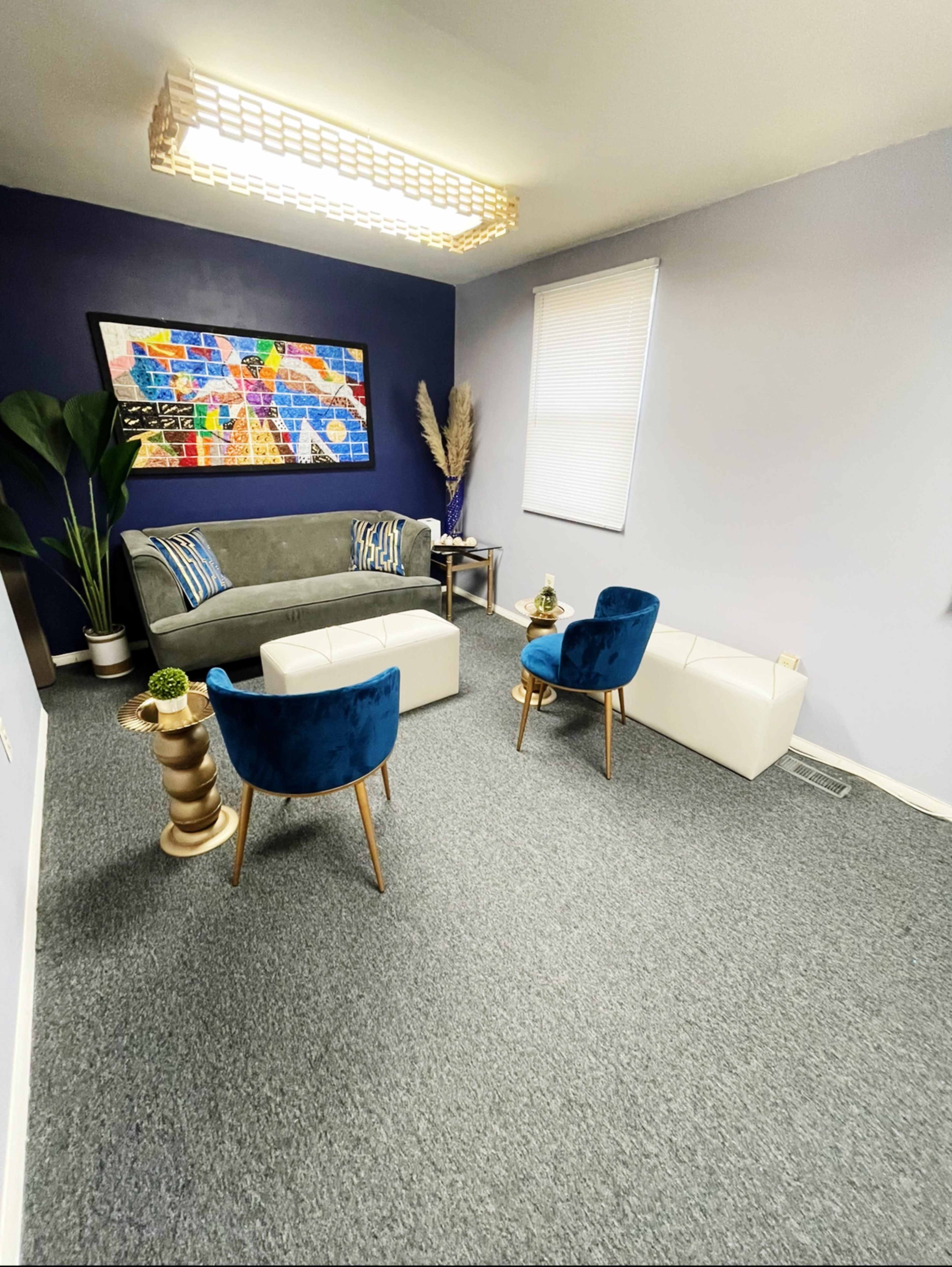 A small, modern sitting area with a gray couch, two blue chairs, a white ottoman, and a large colorful artwork on the wall.