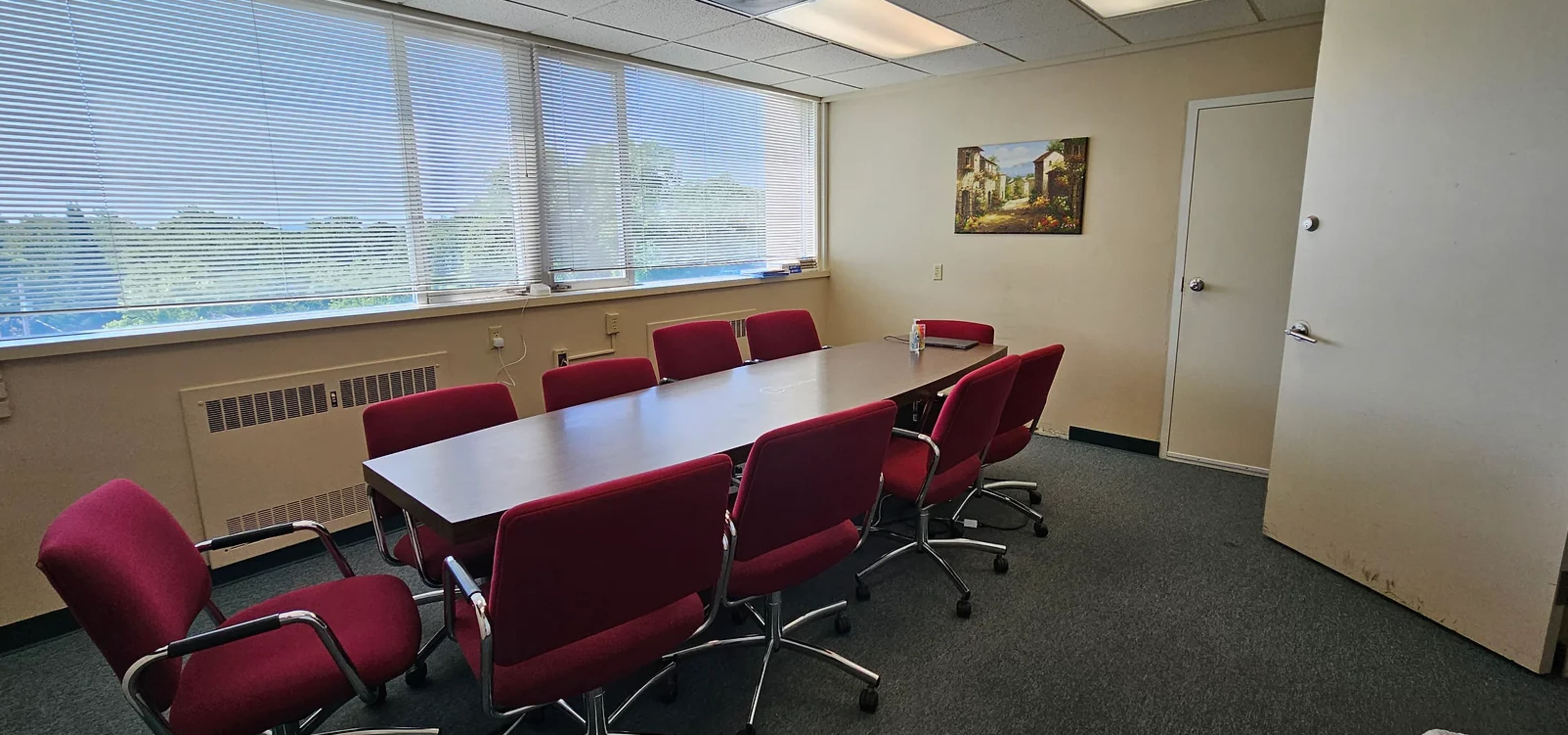 A conference room features a large table surrounded by several red chairs, with windows allowing natural light and a painting on the wall.