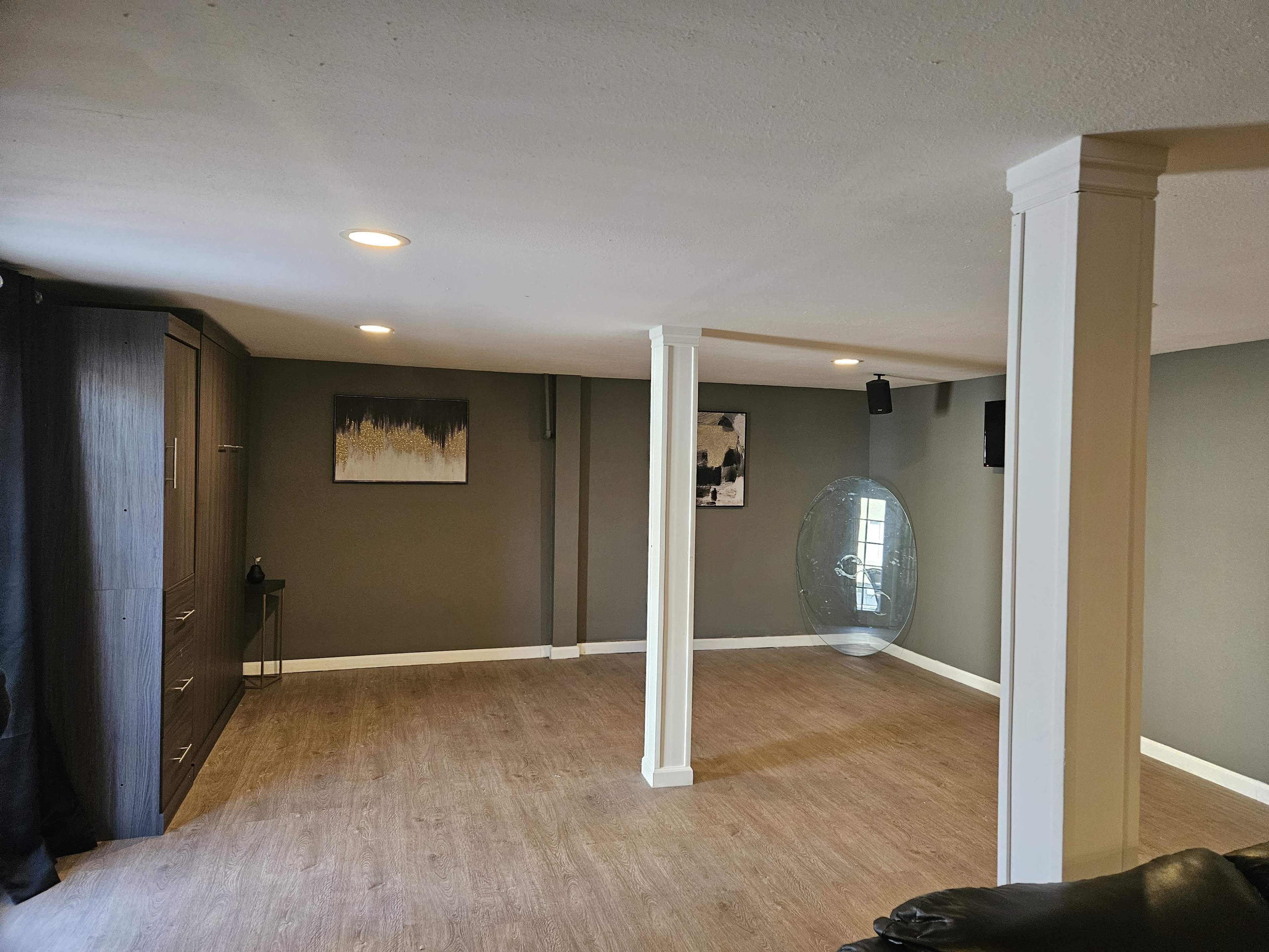 The image shows a spacious room with wood flooring, two support pillars, and minimal furnishings, including a wall-mounted television and abstract wall art.