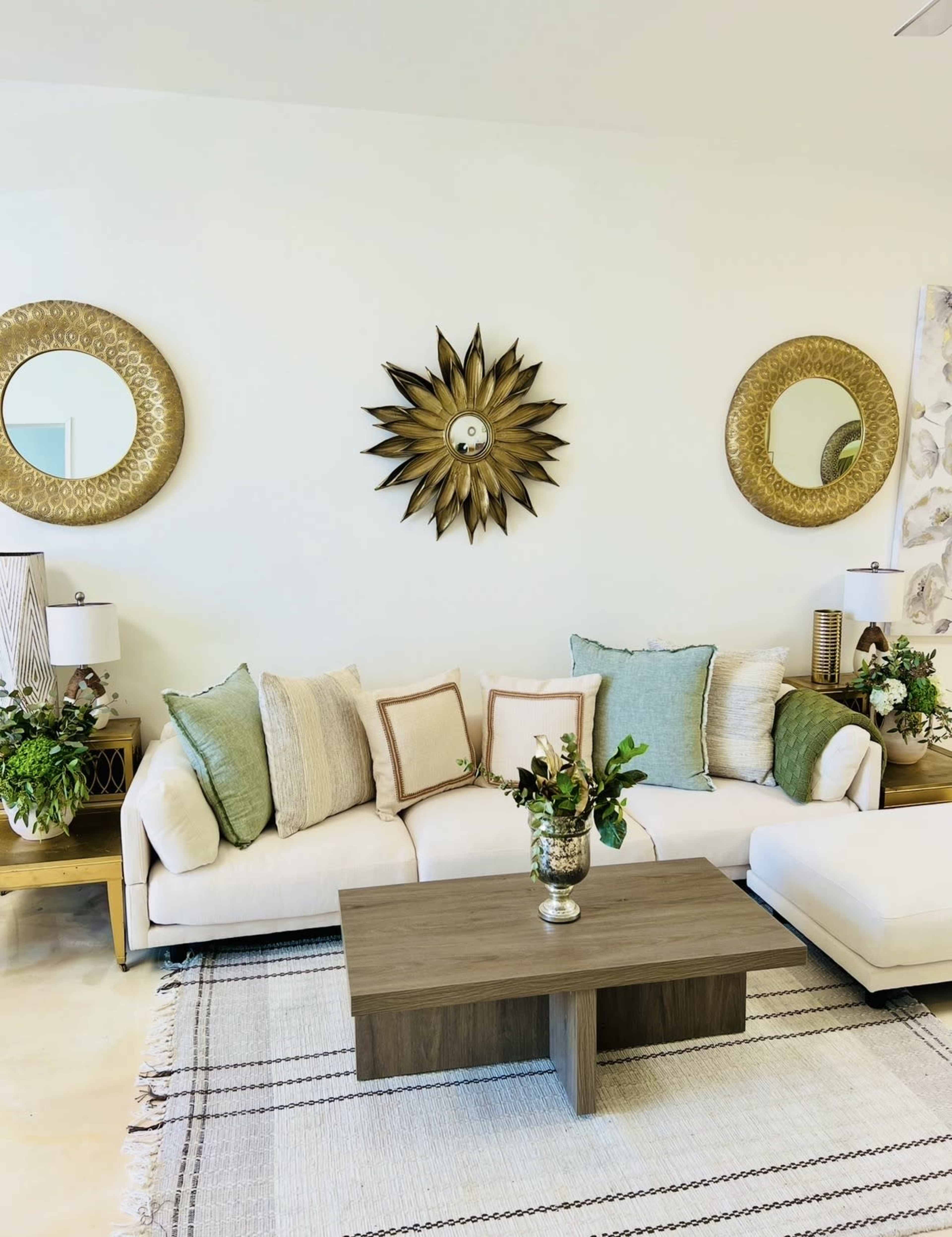 A beige sectional sofa with green and cream pillows is positioned in front of a wooden coffee table on a patterned rug, with decorative mirrors and plants on the wall and side tables.