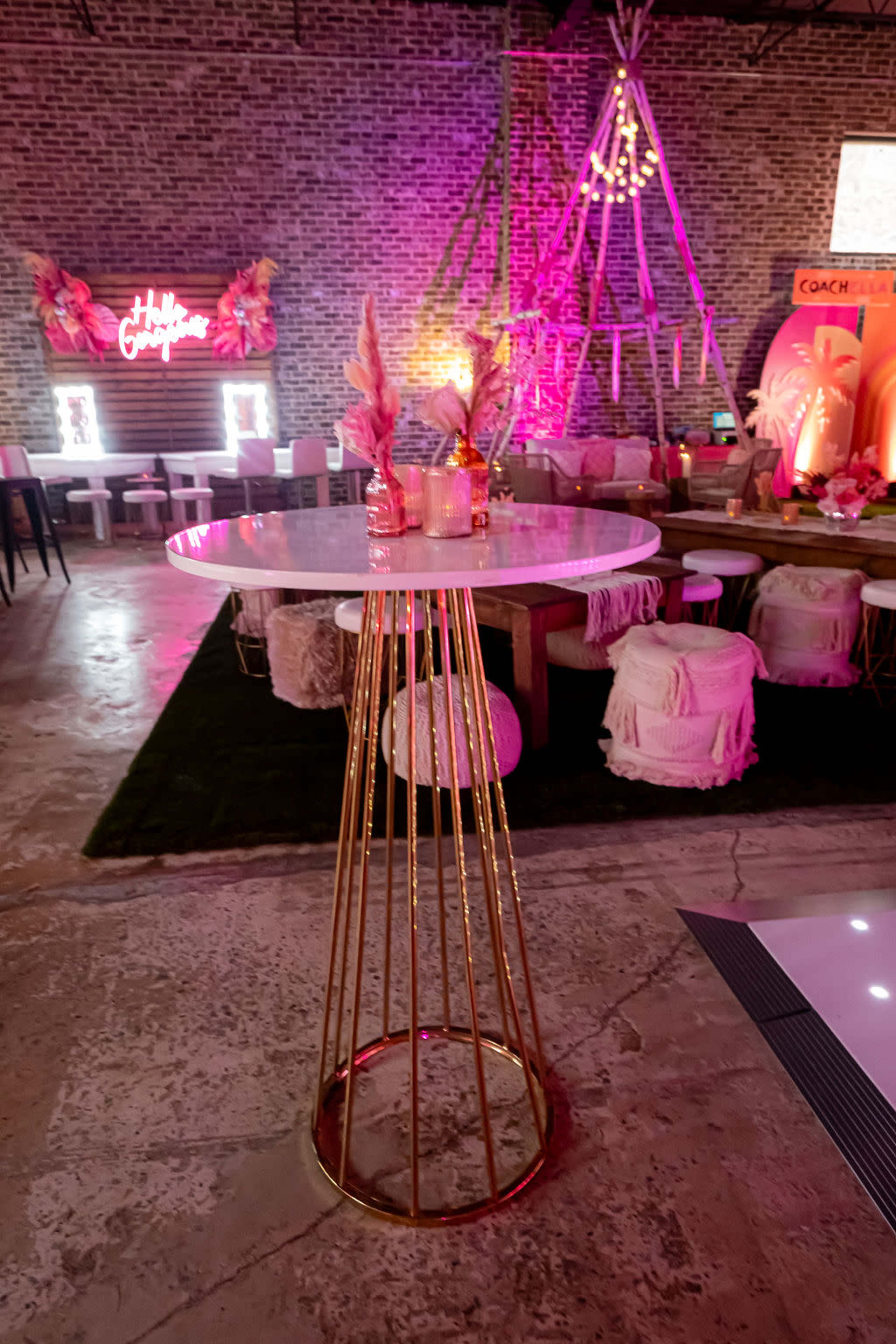 The image shows a modern, tall round table with a gold base, surrounded by a vibrant event space decorated with colorful lights and stylish seating.