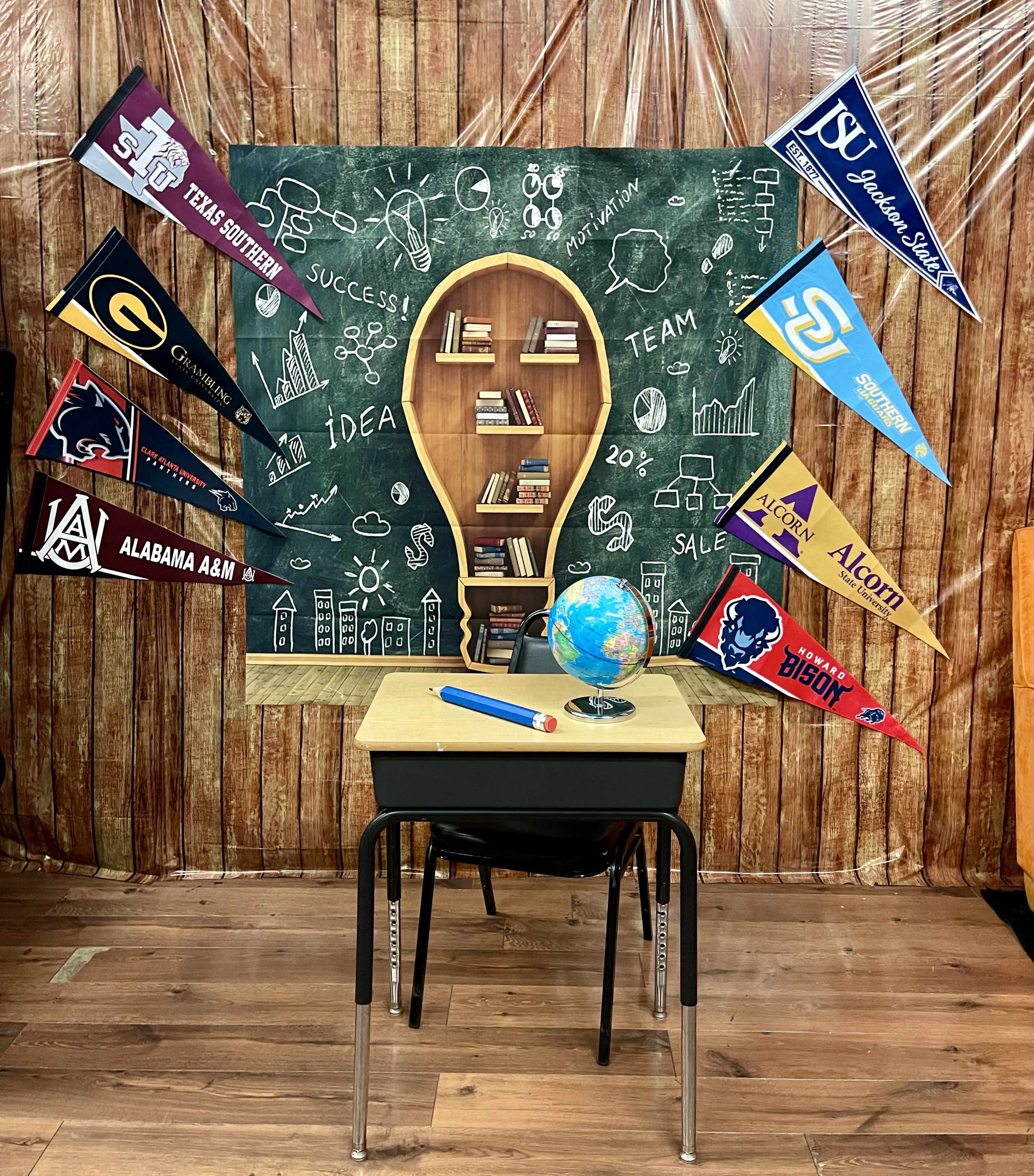 The image shows a classroom setup with a desk in front of a chalkboard displaying a light bulb graphic, surrounded by pennants from different universities.
