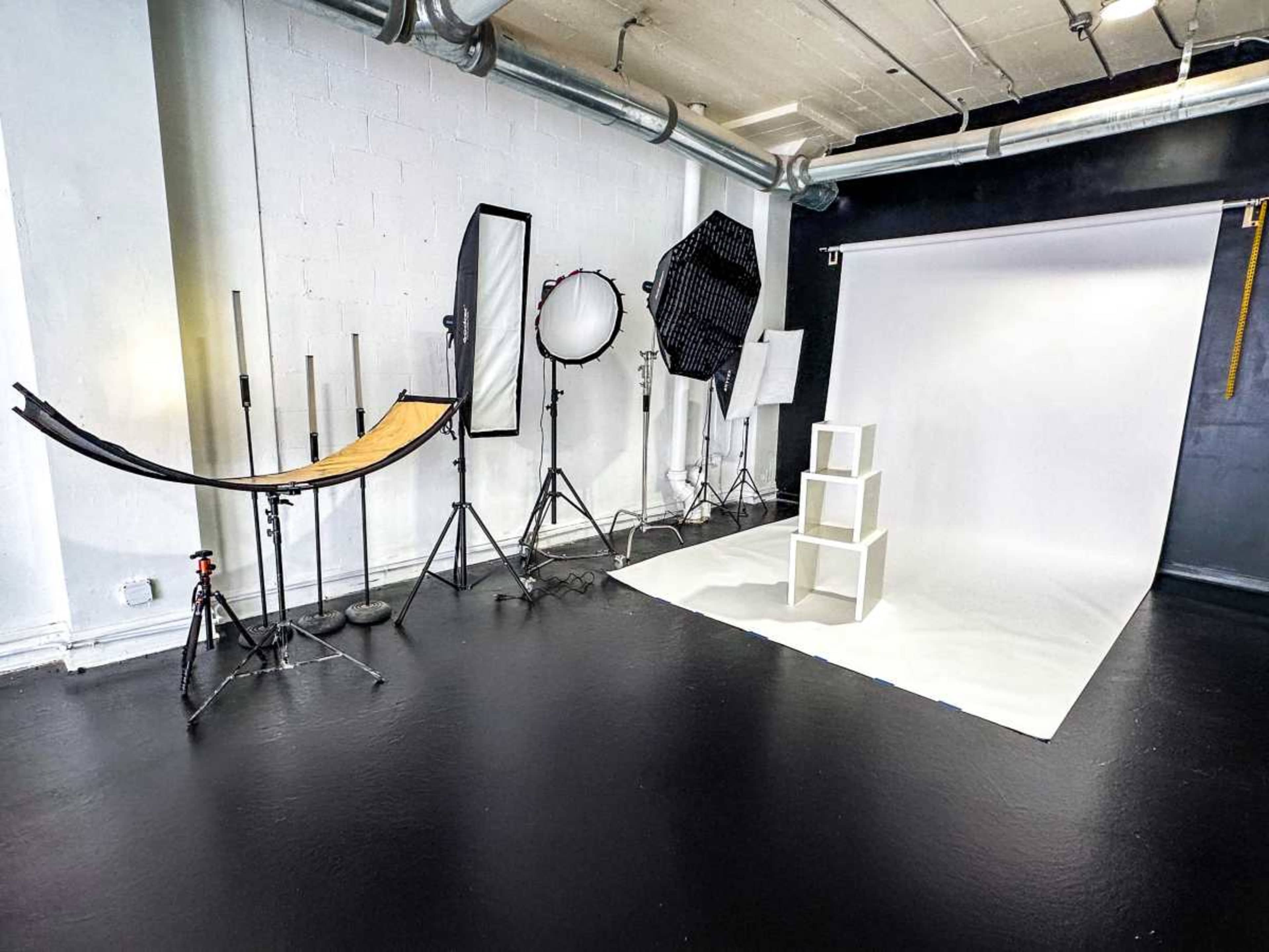 The image depicts a photography studio equipped with various lighting setups and a backdrop, featuring a stack of three white cubes on a white floor.