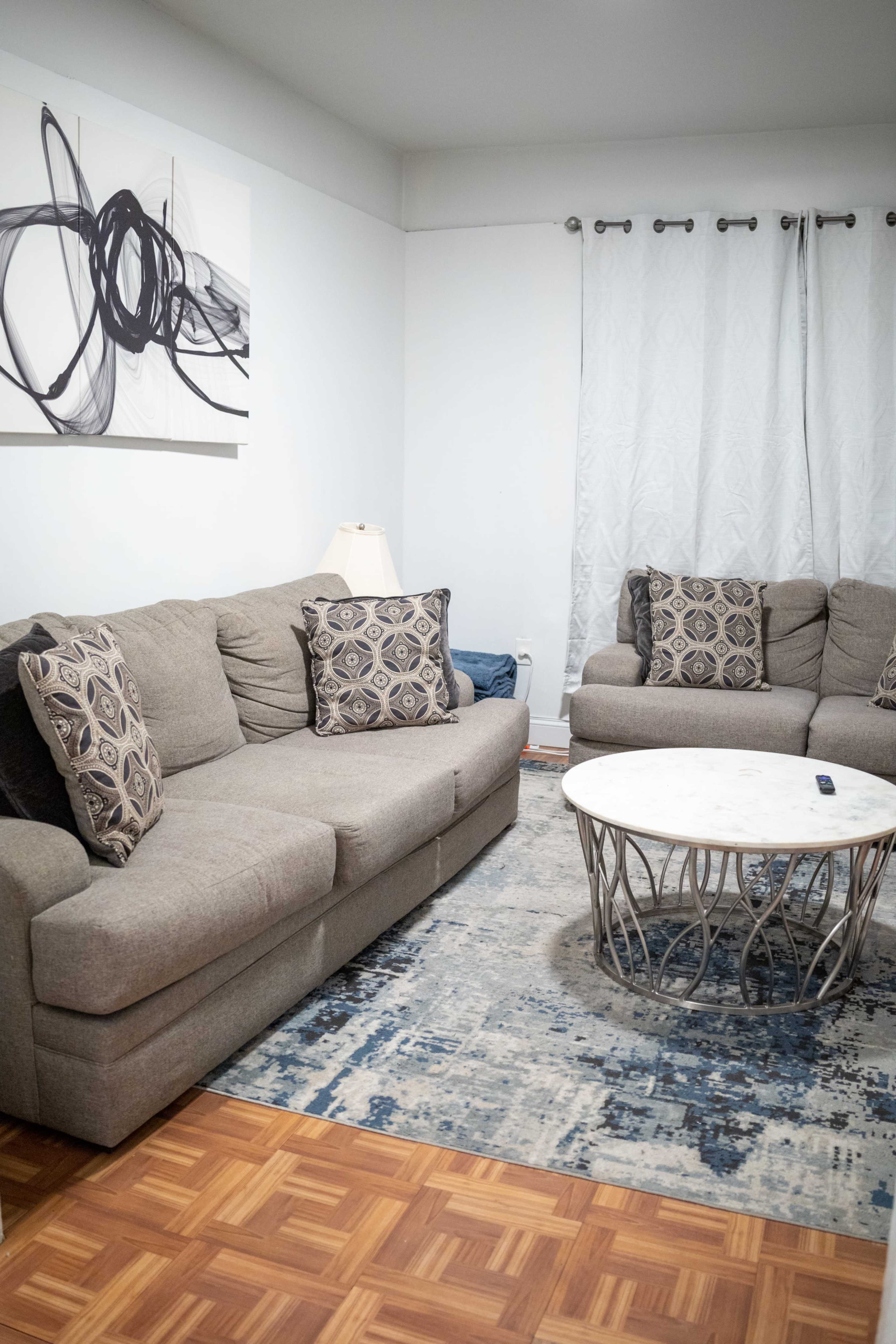 A living room features a gray sectional sofa and matching chairs, with decorative pillows, a round marble coffee table, and a light-colored area rug on a wooden floor.