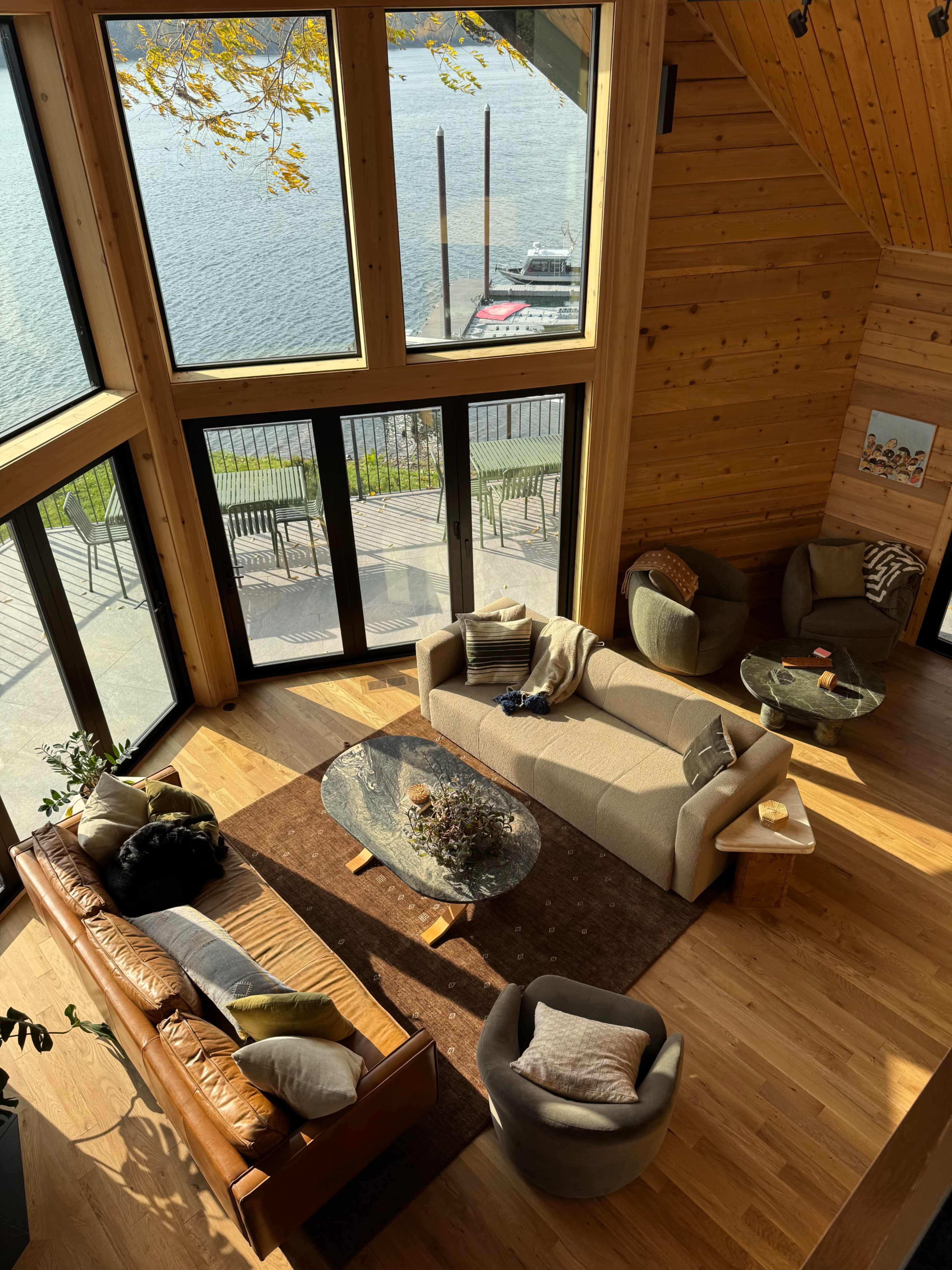 A spacious living area features large windows overlooking a lake, furnished with a combination of sofas, armchairs, and a glass coffee table on a wooden floor.