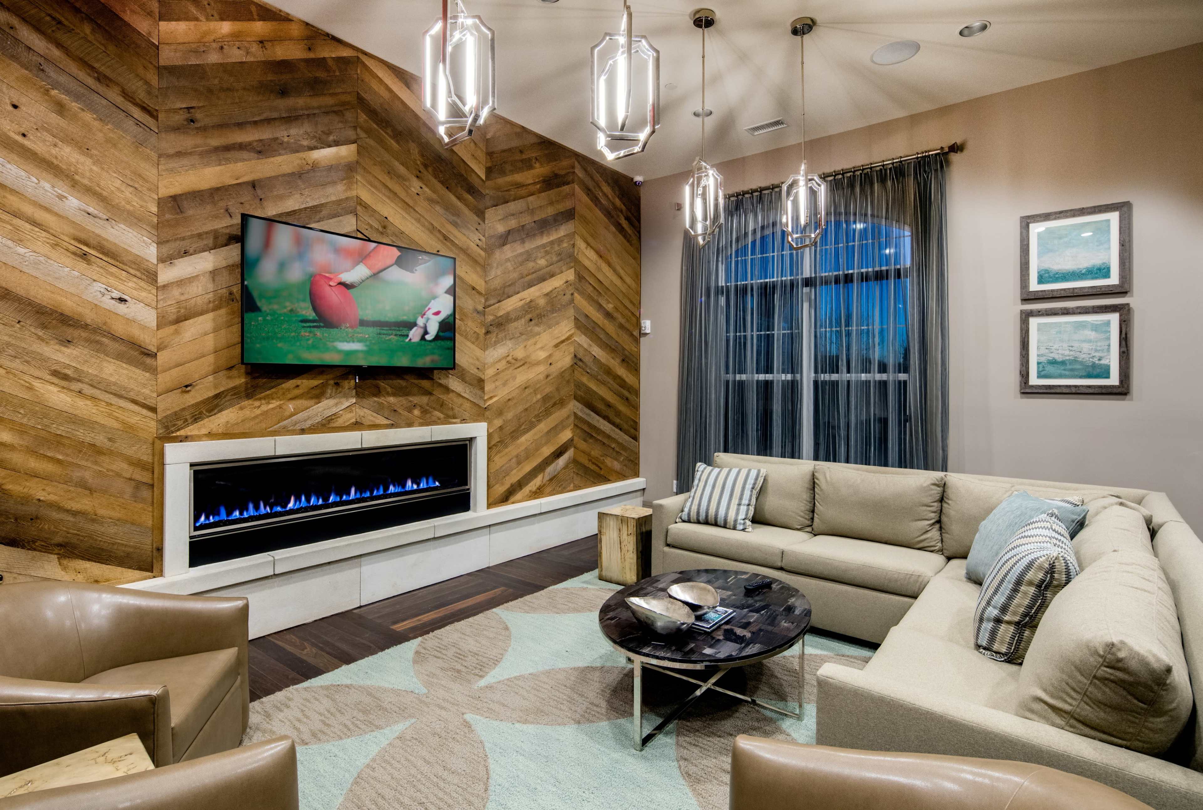A modern living room featuring a sectional sofa, armchairs, a round coffee table, a wall-mounted television, and a fireplace beneath the TV, all set against a backdrop of wood-paneled walls.