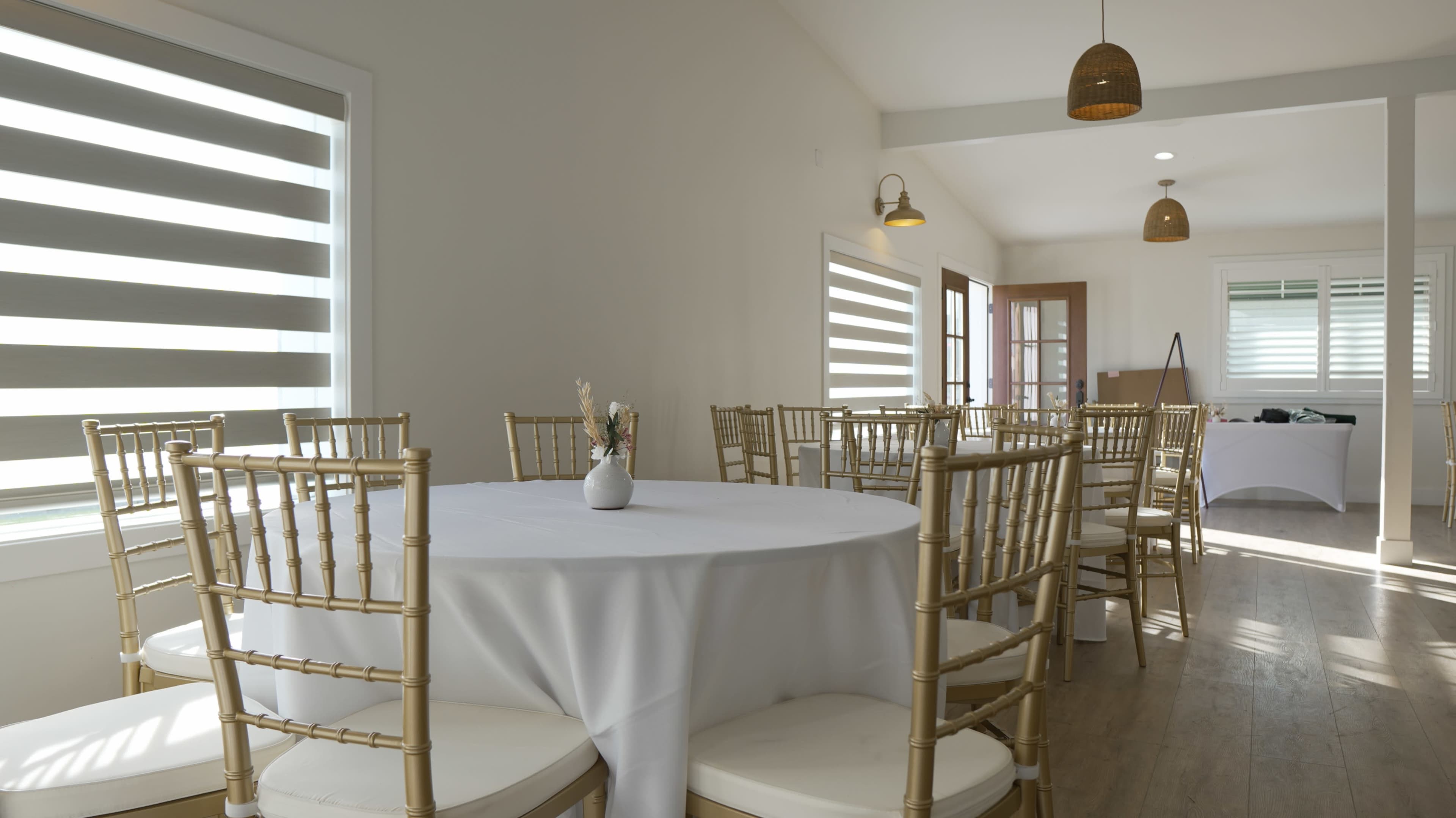 A bright, minimalist room features round tables with white tablecloths and gold chiavari chairs, illuminated by natural light through striped windows.