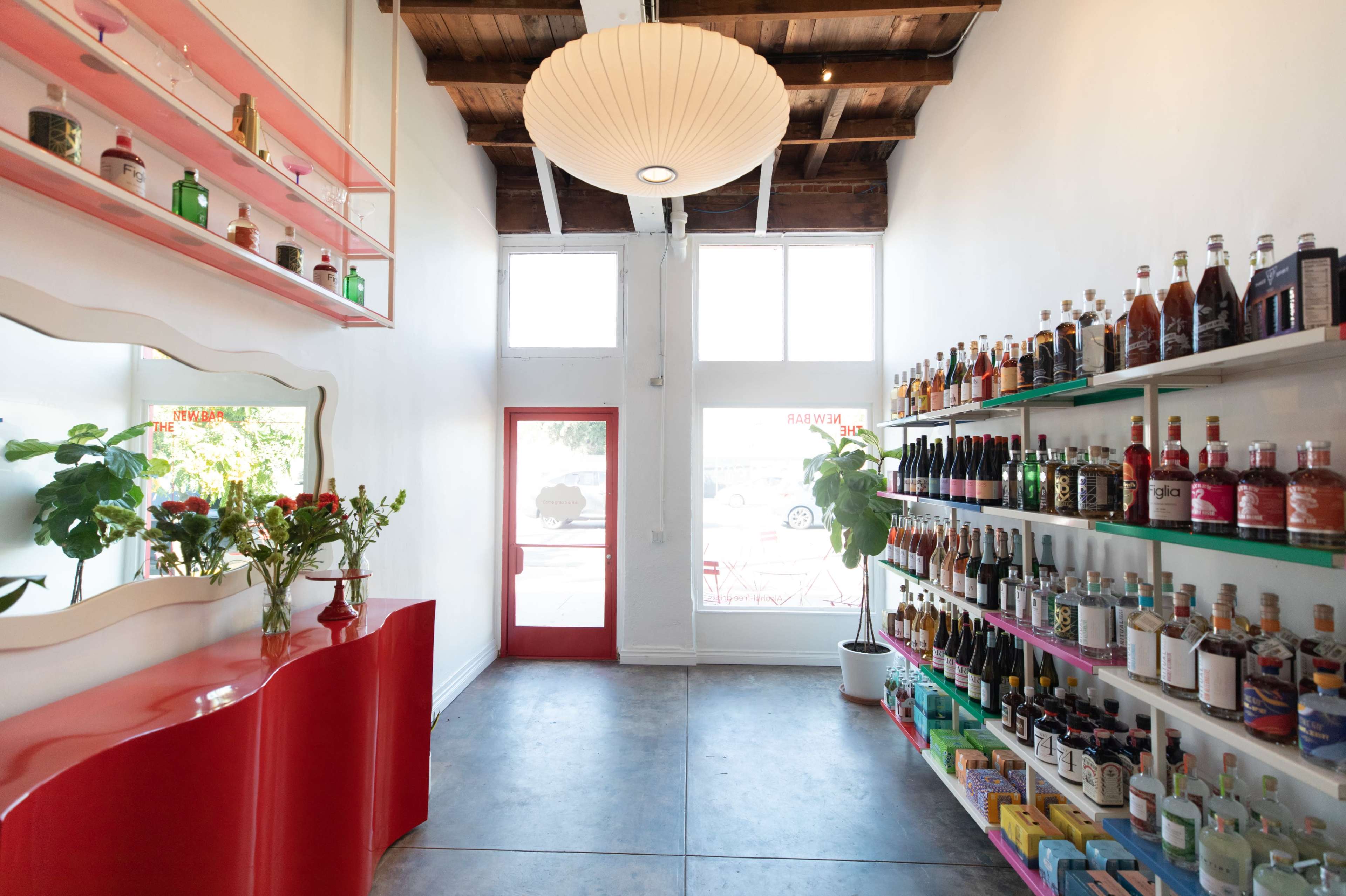 The interior of a modern store features a red display counter and shelves filled with various bottles, alongside a large pendant light and windows letting in natural light.