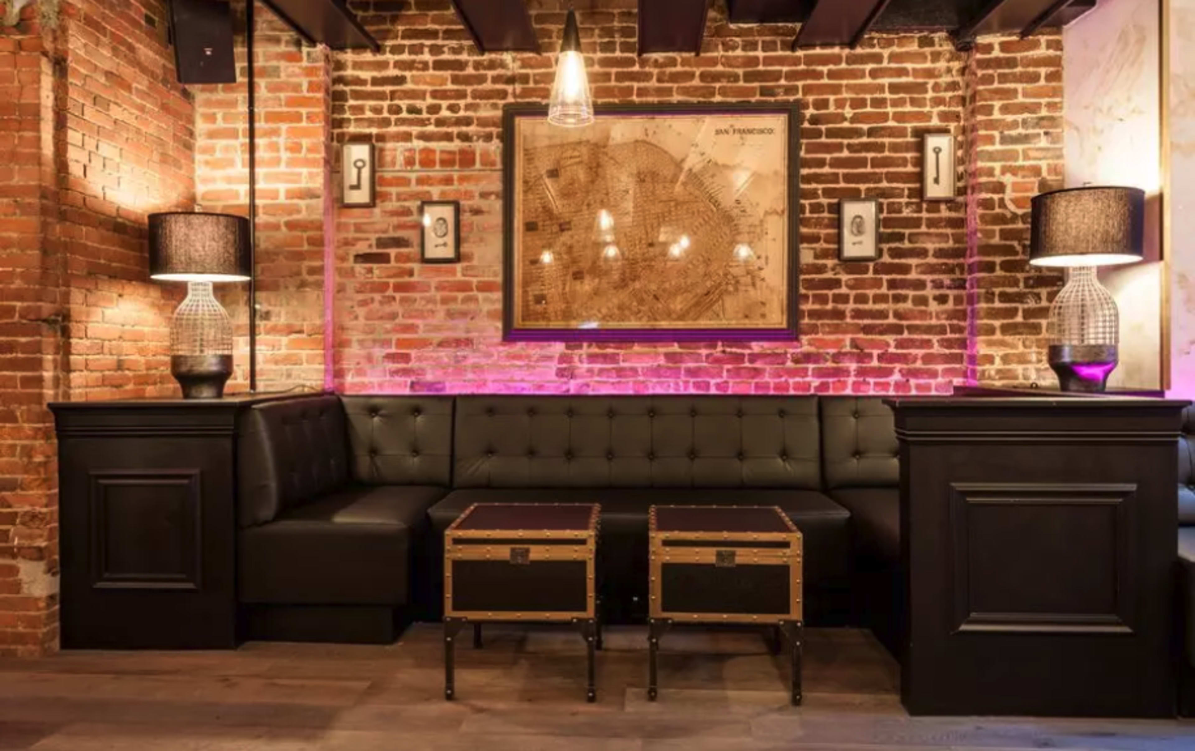 The image shows a cozy seating area in a restaurant featuring a tufted bench along a brick wall, illuminated by soft lighting, with a vintage map framed on the wall above two wooden tables.