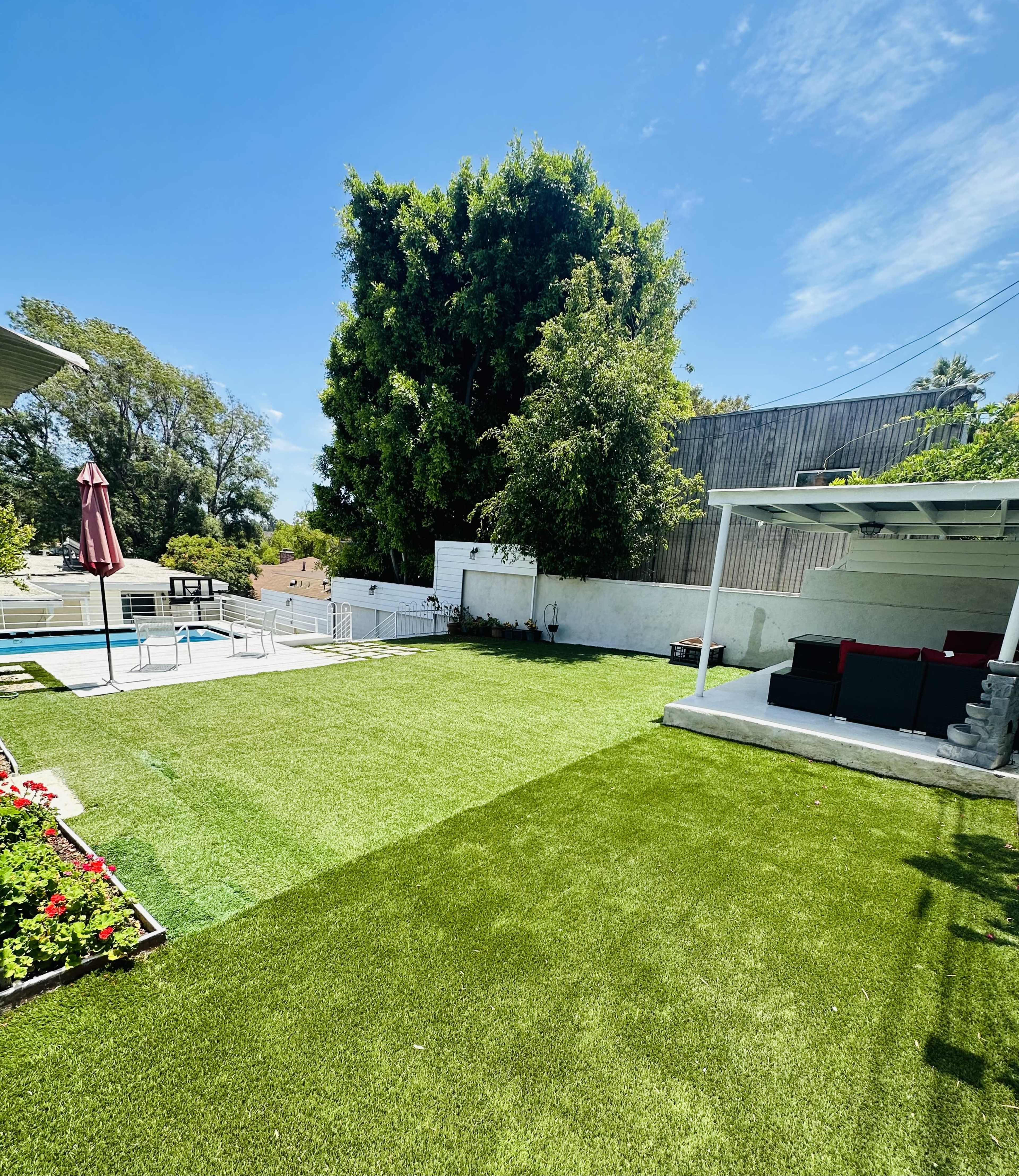 A well-maintained backyard with artificial grass, a swimming pool in the distance, and a seating area under a covered patio.