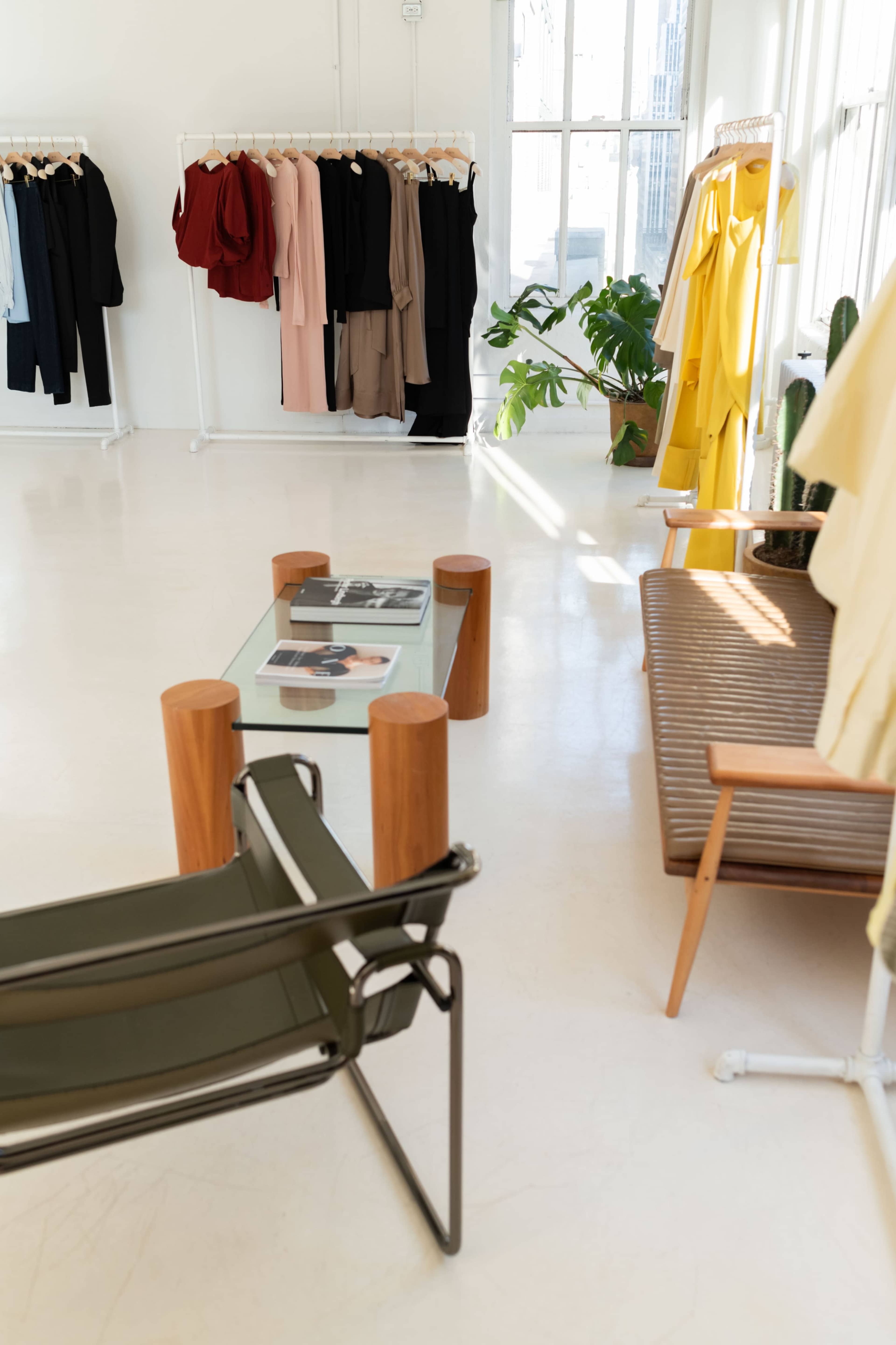 The image shows a bright boutique with hanging clothing displays on racks, a glass coffee table, a wooden bench, and potted plants.