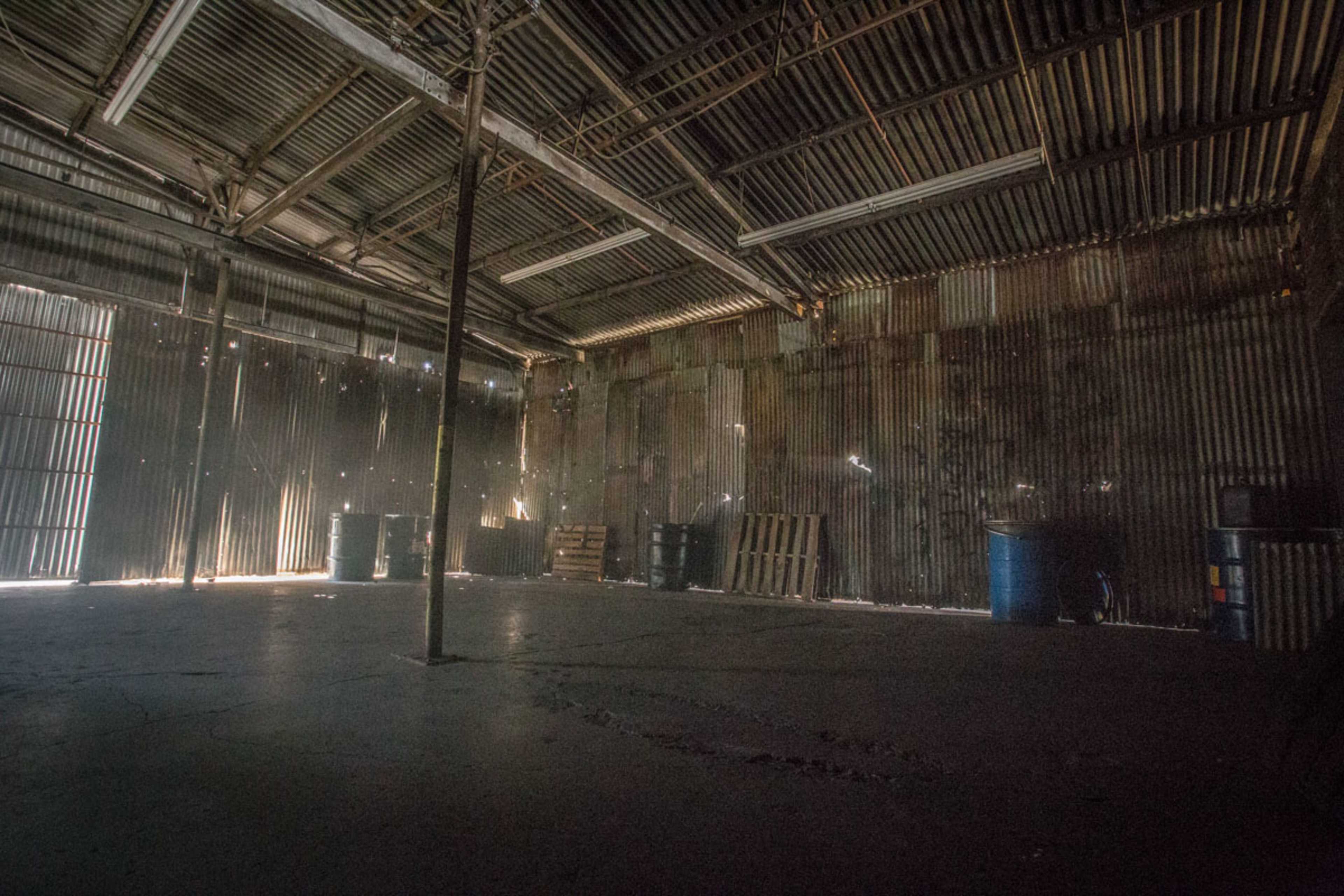 The image shows an empty industrial warehouse with metal walls, a concrete floor, and several barrels positioned against the walls.