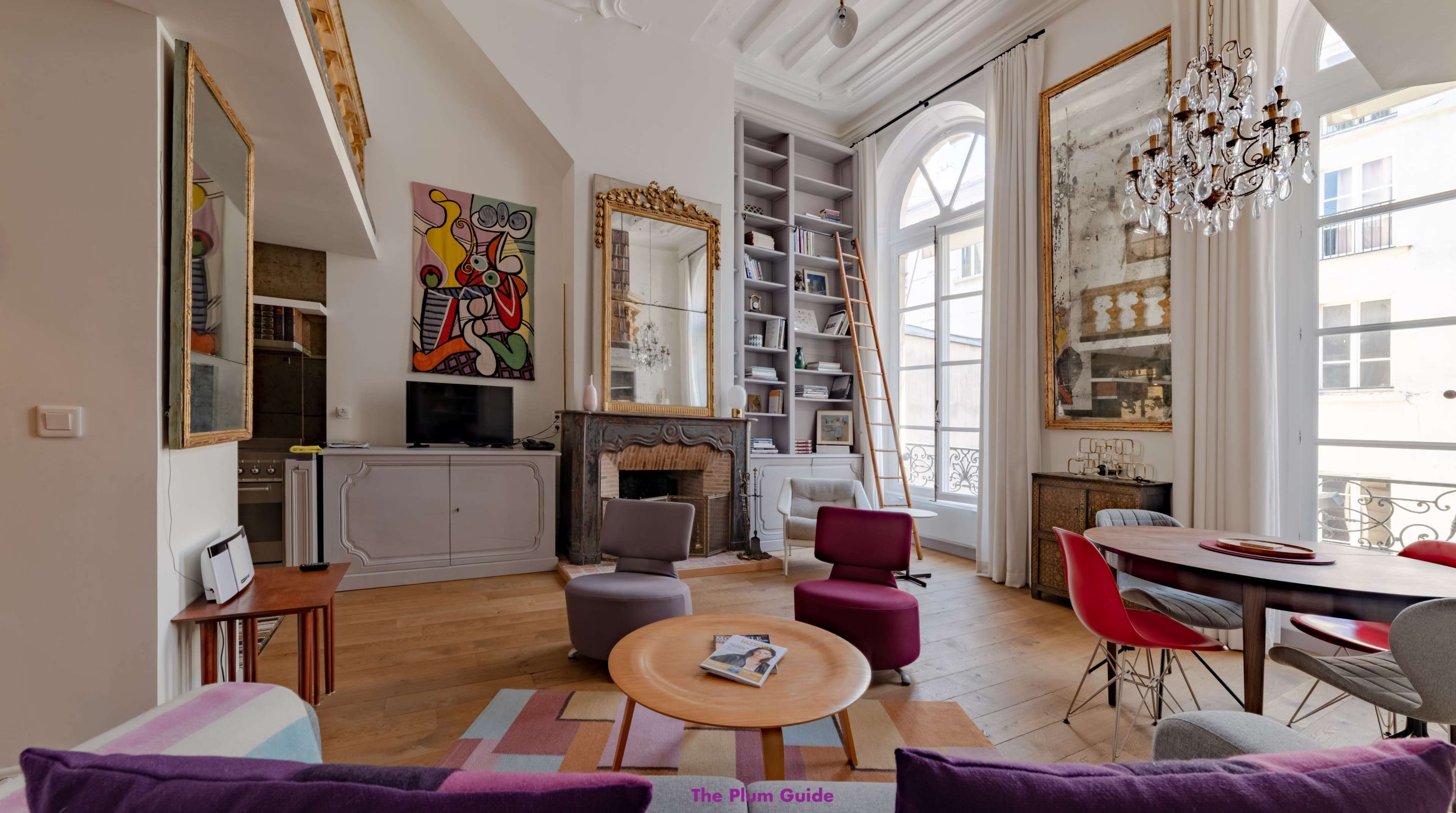 A stylish living room features large windows, a modern art piece, mirrored walls, and a blend of colorful furniture, including a circular coffee table and a dining area.