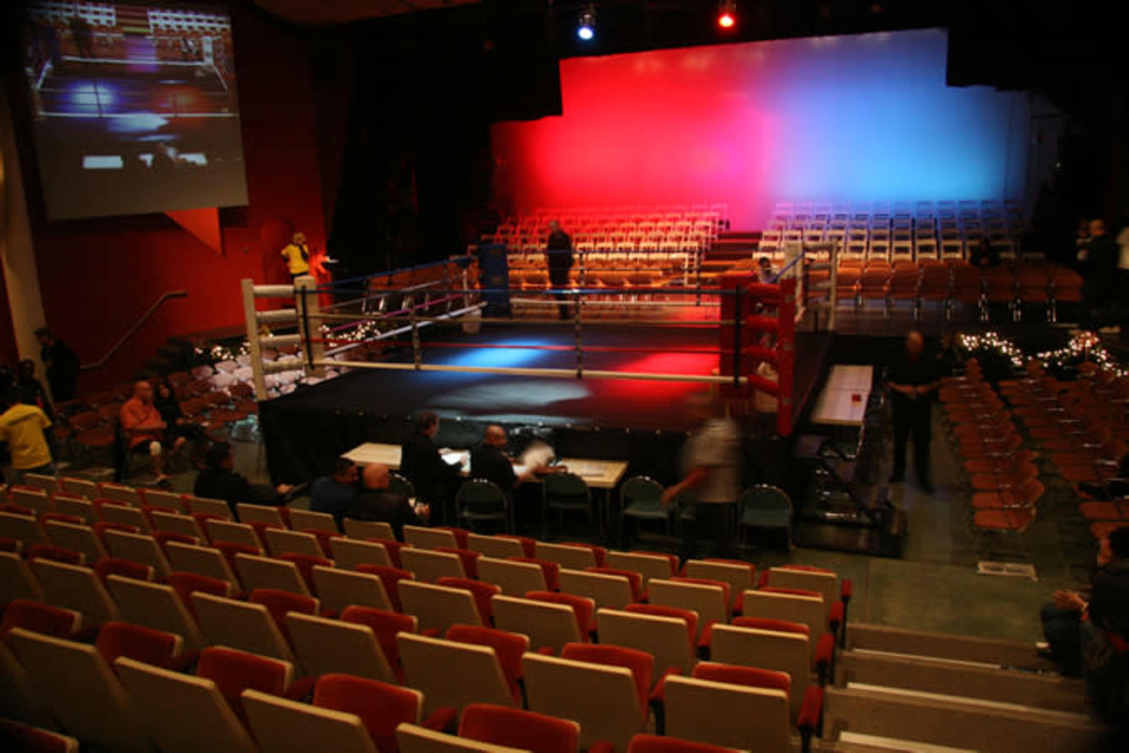 The image shows a boxing ring set up in a theater-like venue with empty seats and a large screen displaying a boxing match in the background.