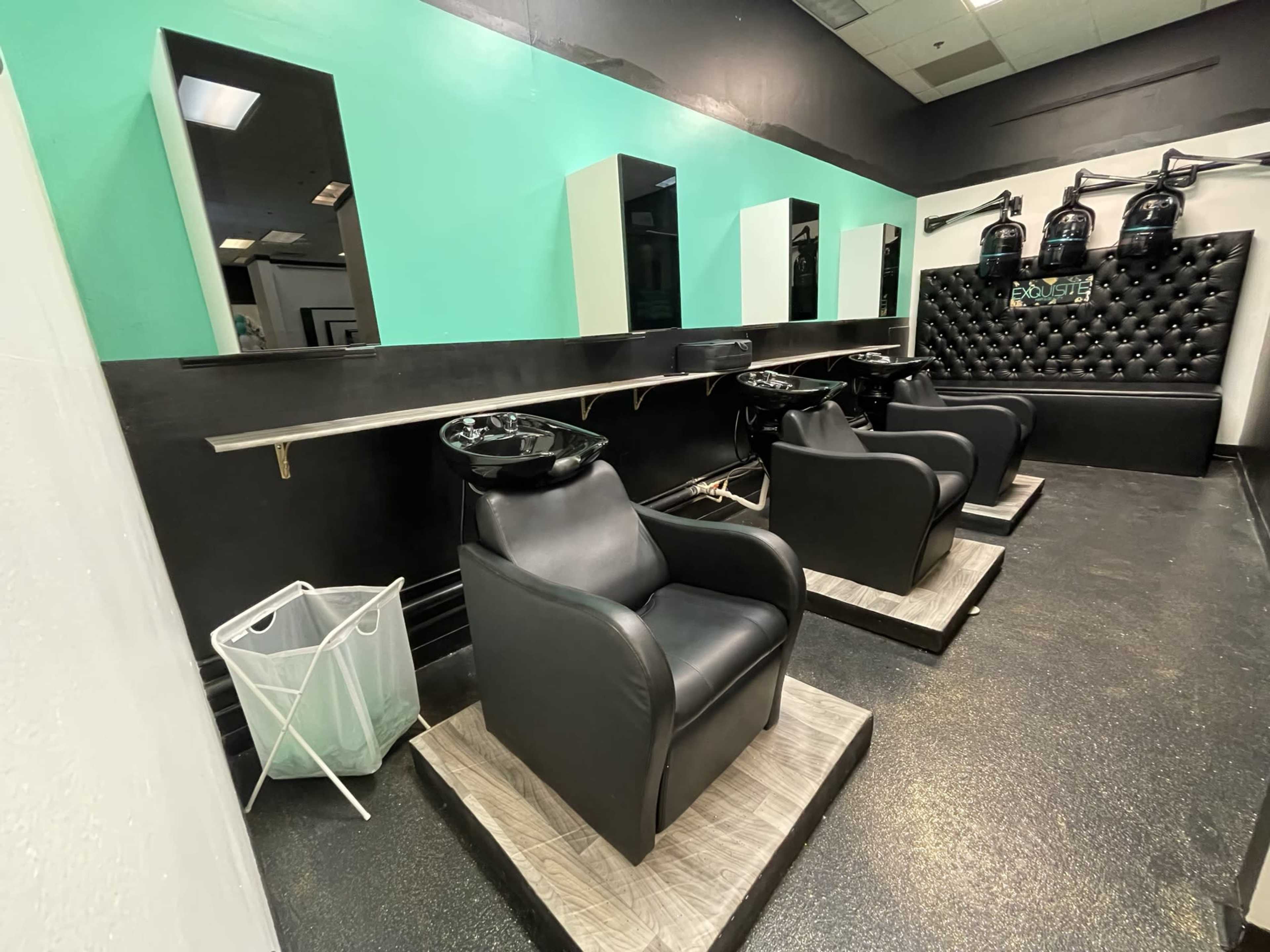 A hair salon interior featuring two black chairs with shampoo sinks, mirrors on the wall, and a black tufted section with hair dryers.