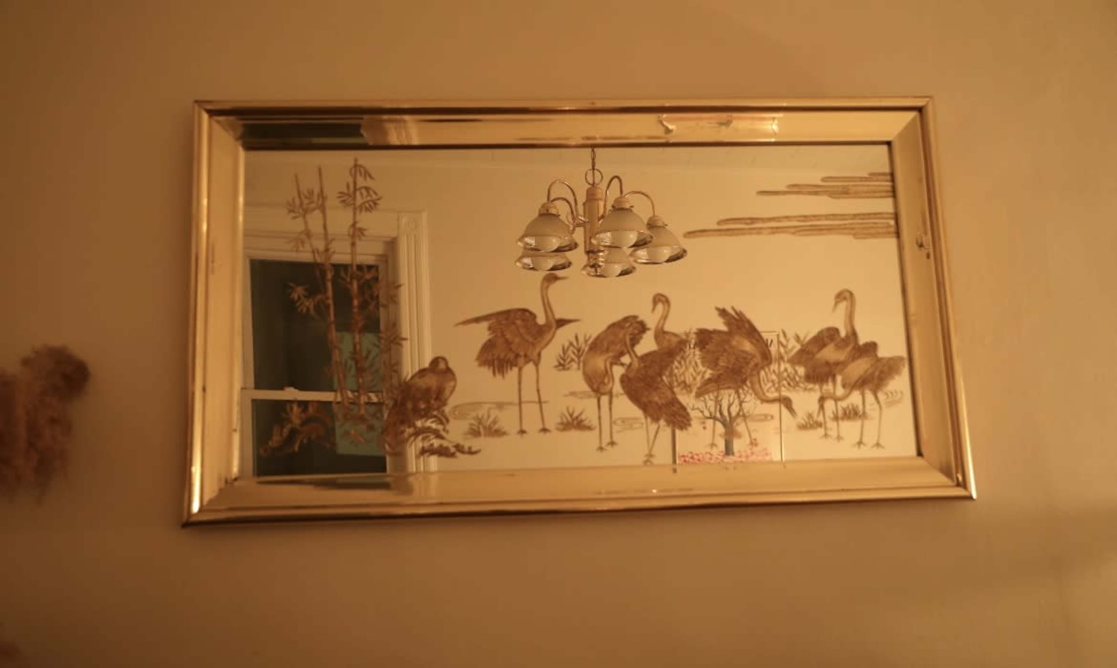 A framed mirror hangs on the wall, reflecting a design of cranes and vegetation, with a light fixture visible above it.
