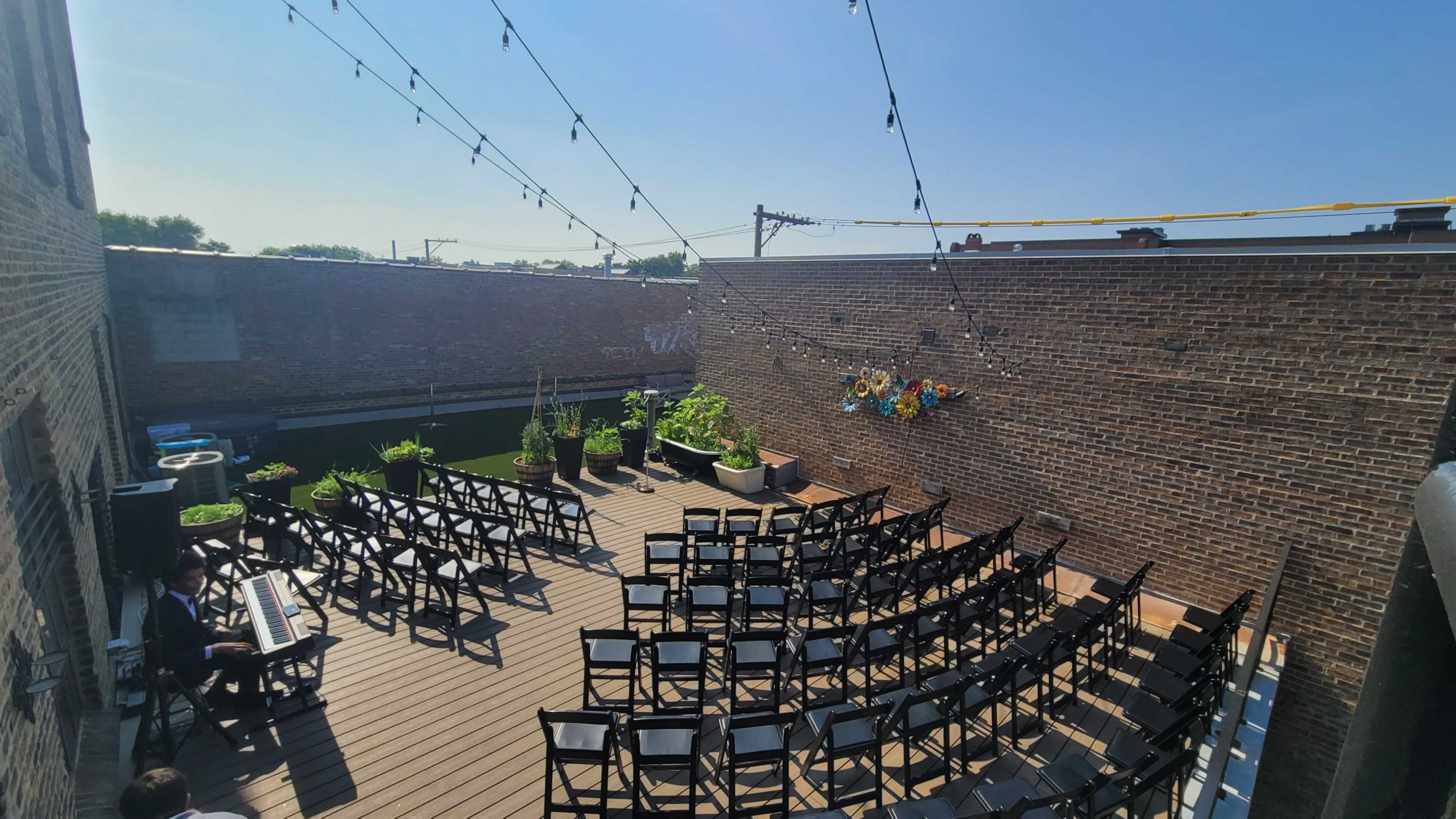 A rooftop event space features rows of black chairs arranged on a wooden deck, with string lights overhead and potted plants lining the edges.