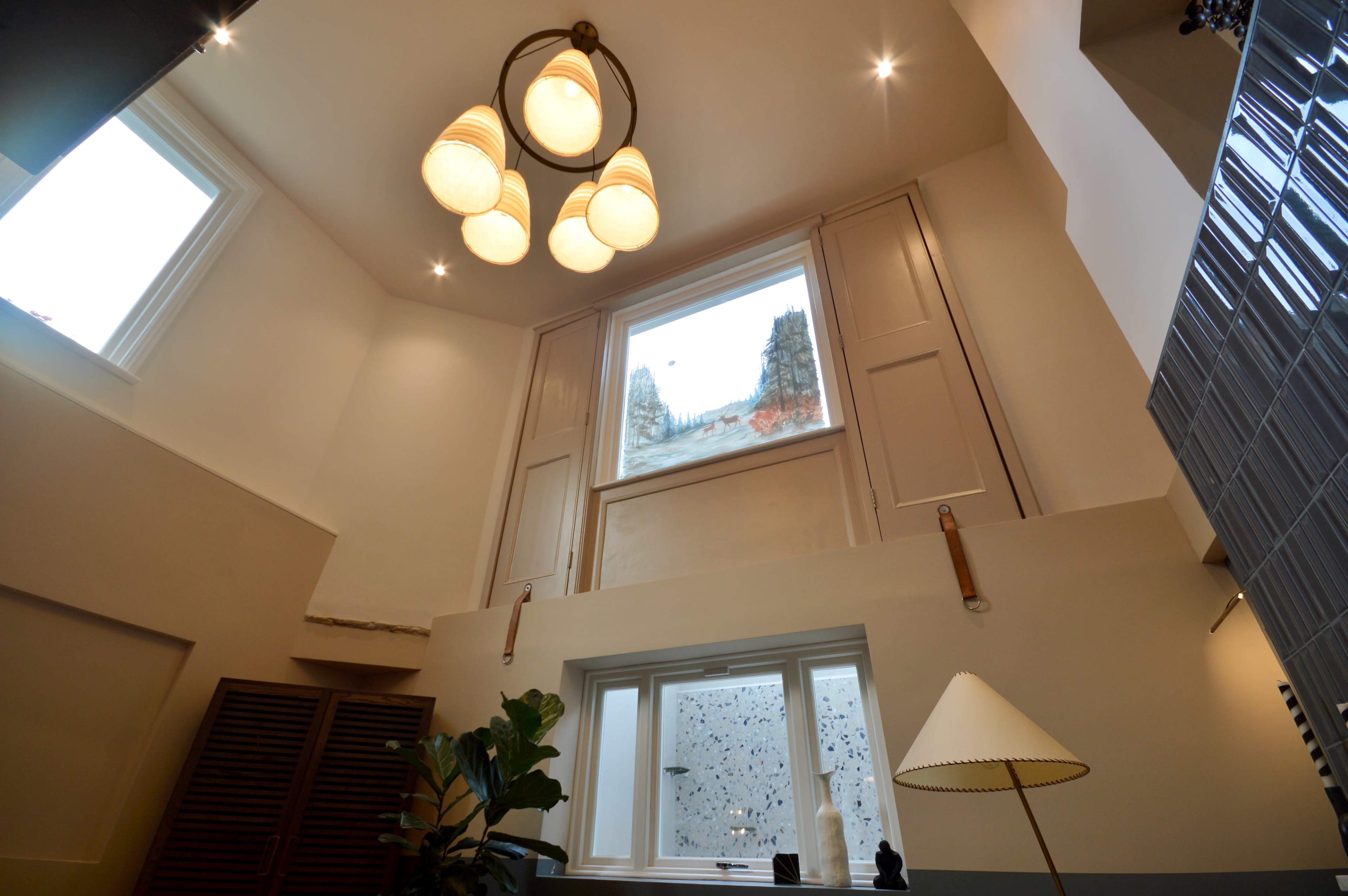The image shows a modern interior space with high ceilings, a large window, decorative light fixtures, and a mix of architectural elements.