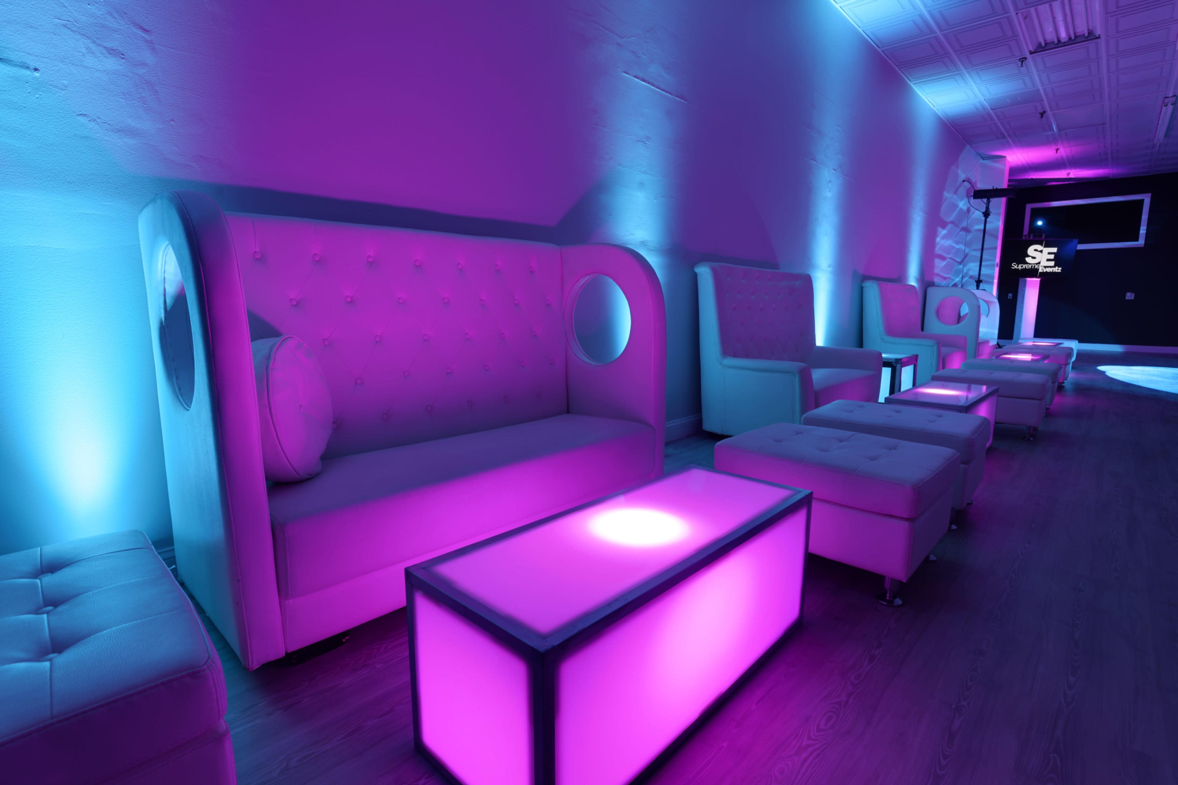 The image shows a modern lounge area with purple and blue lighting, featuring stylish couches and illuminated coffee tables.