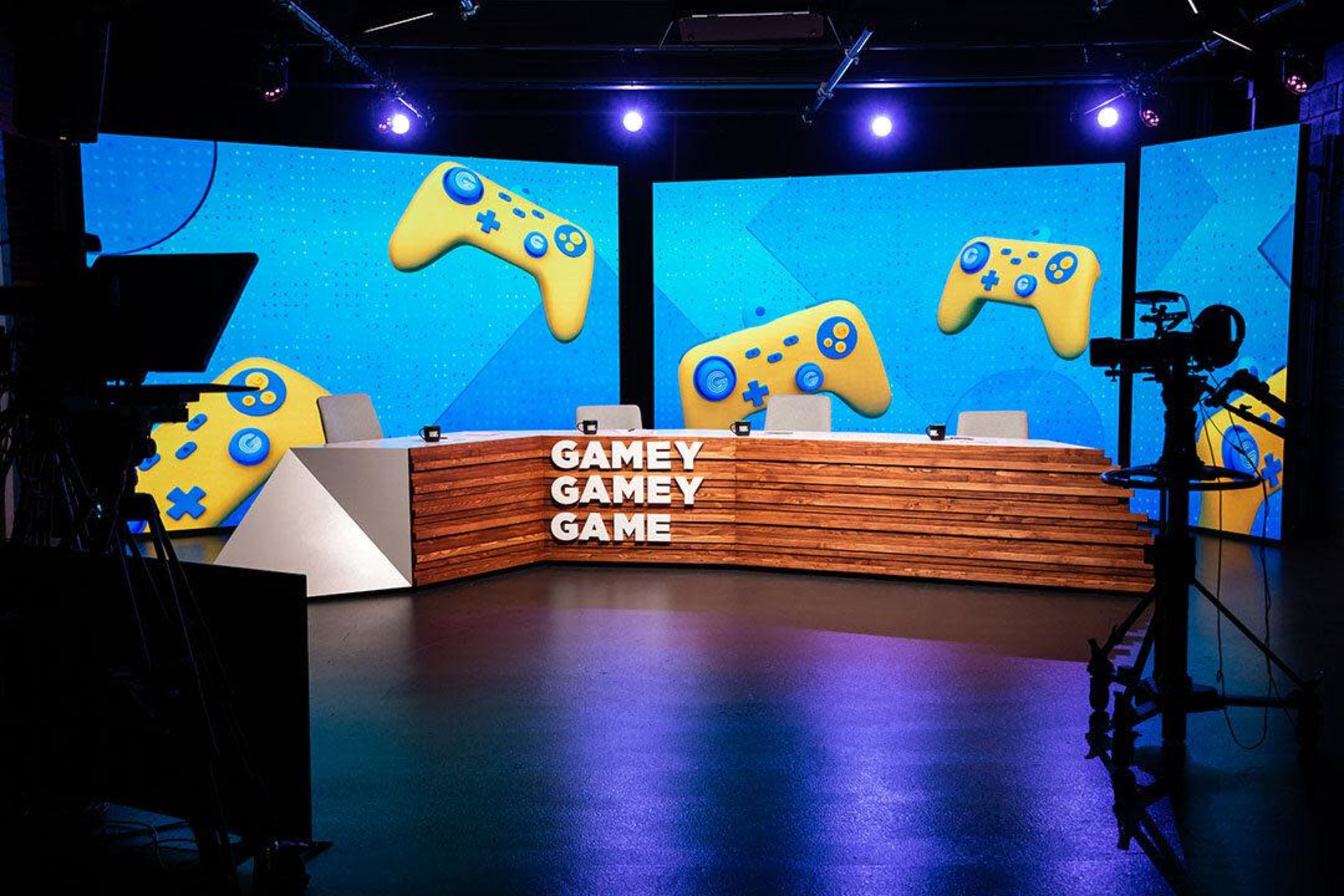A television studio set features a wooden desk with the words "GAMEY GAMEY GAME" and bright blue backgrounds displaying yellow gaming controllers.