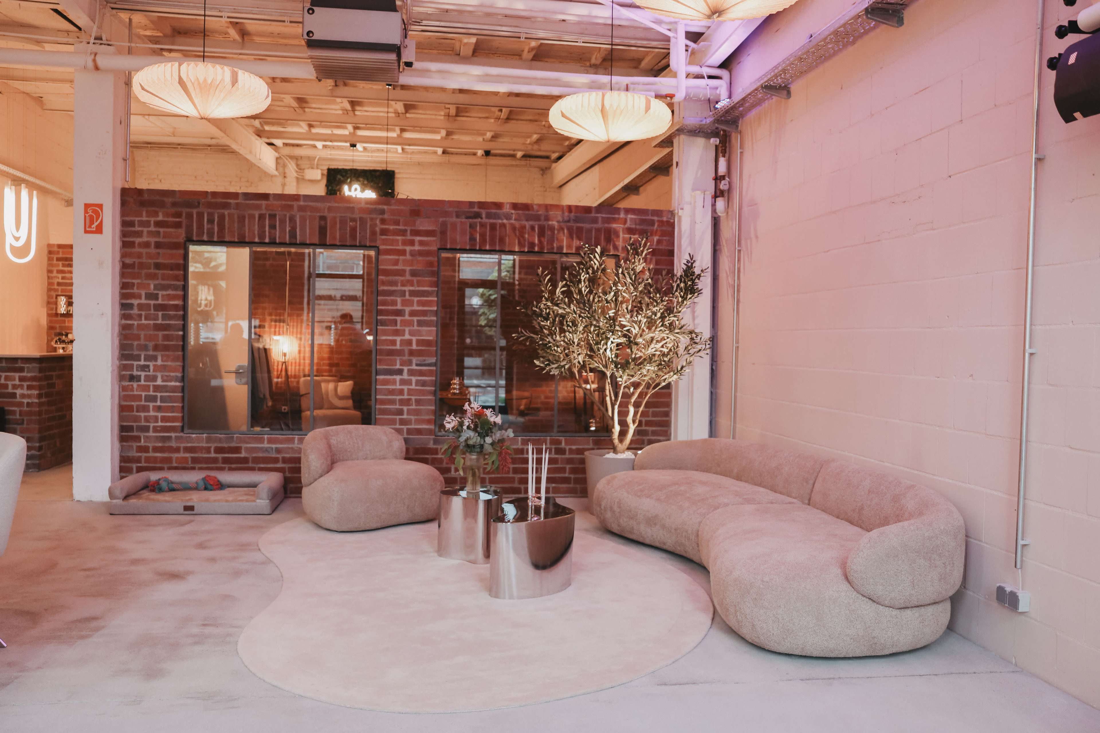 The image shows a modern, minimalist lounge area featuring a curved beige sofa, decorative potted plants, and contemporary coffee tables in a spacious, well-lit room with exposed brick walls.