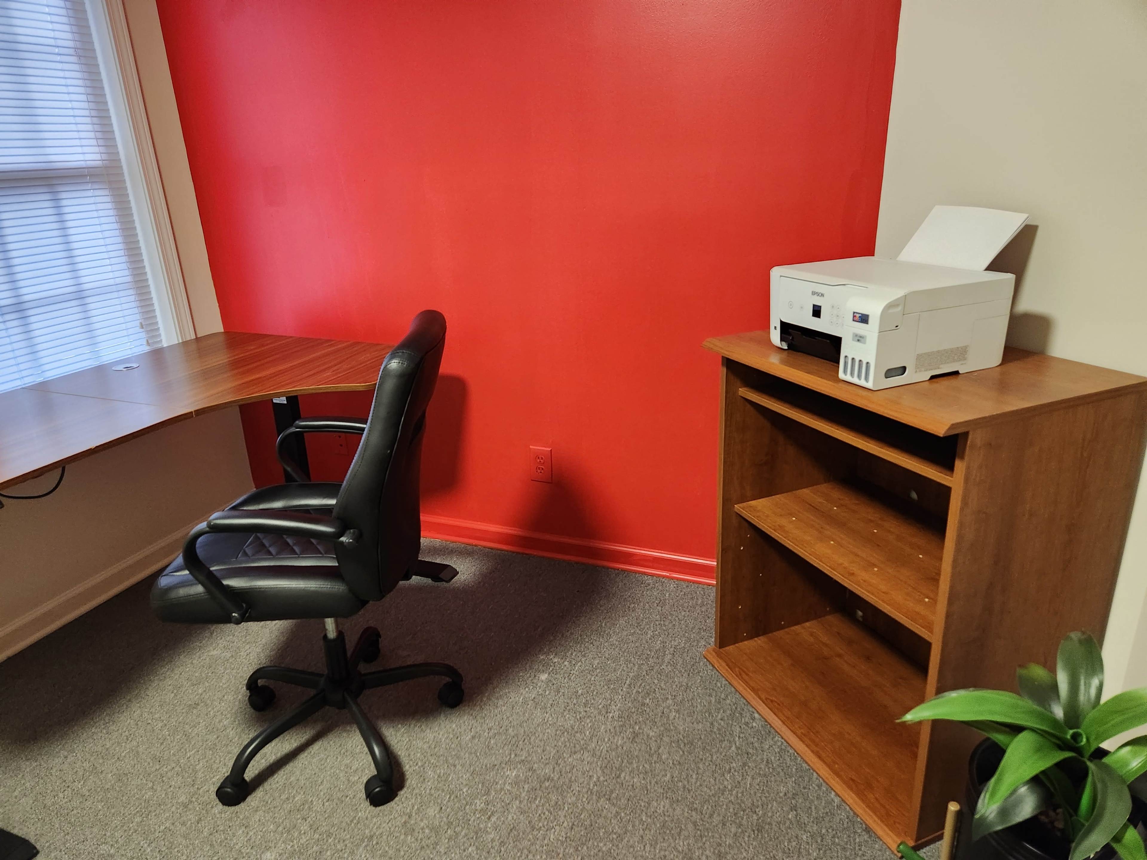 A corner of an office with a wooden desk and a black office chair facing a red wall, alongside a wooden shelf and a printer.