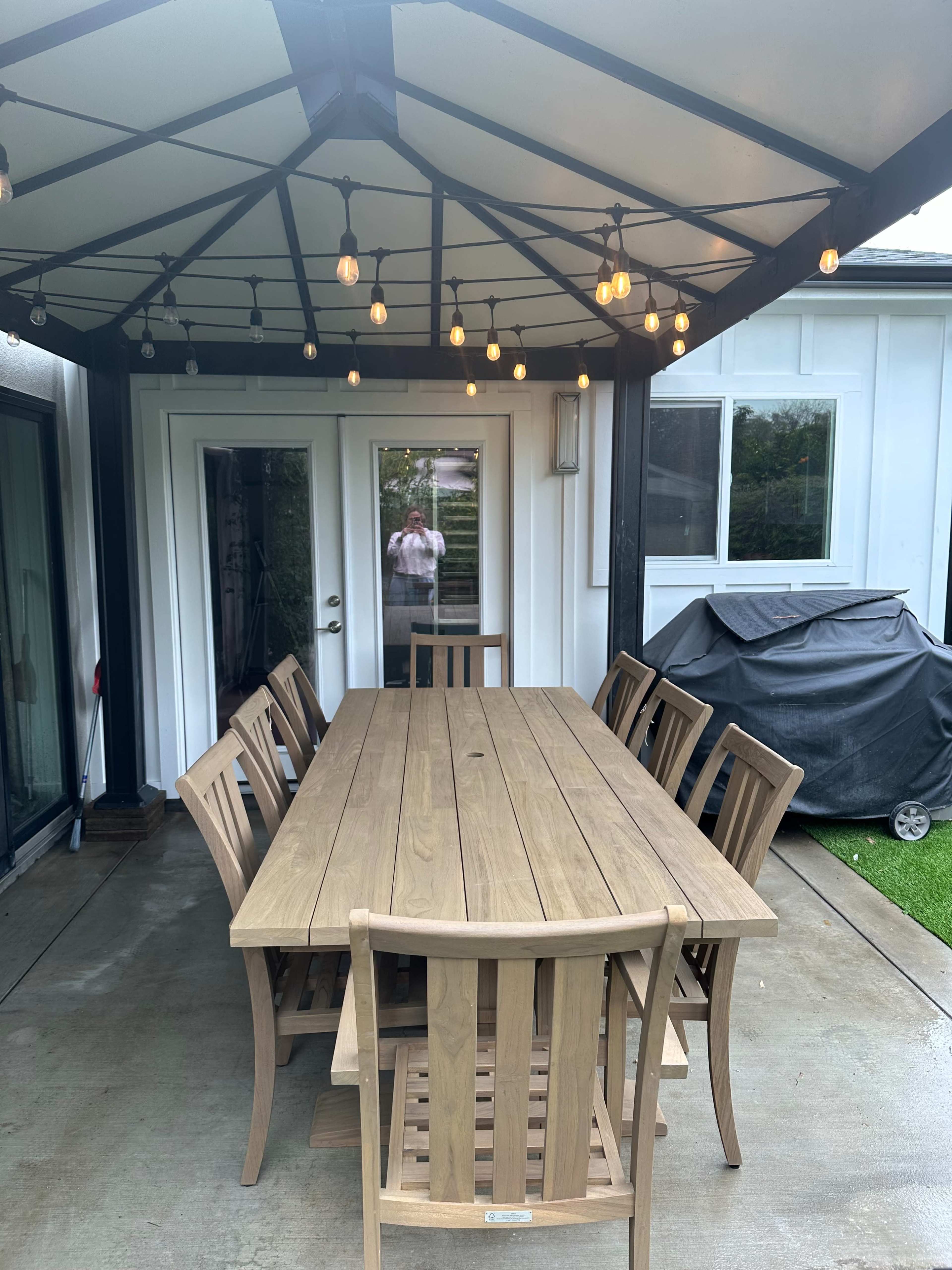 A spacious wooden dining table with matching chairs is set under a canopy with decorative lights, adjacent to a patio door and a barbecue grill.