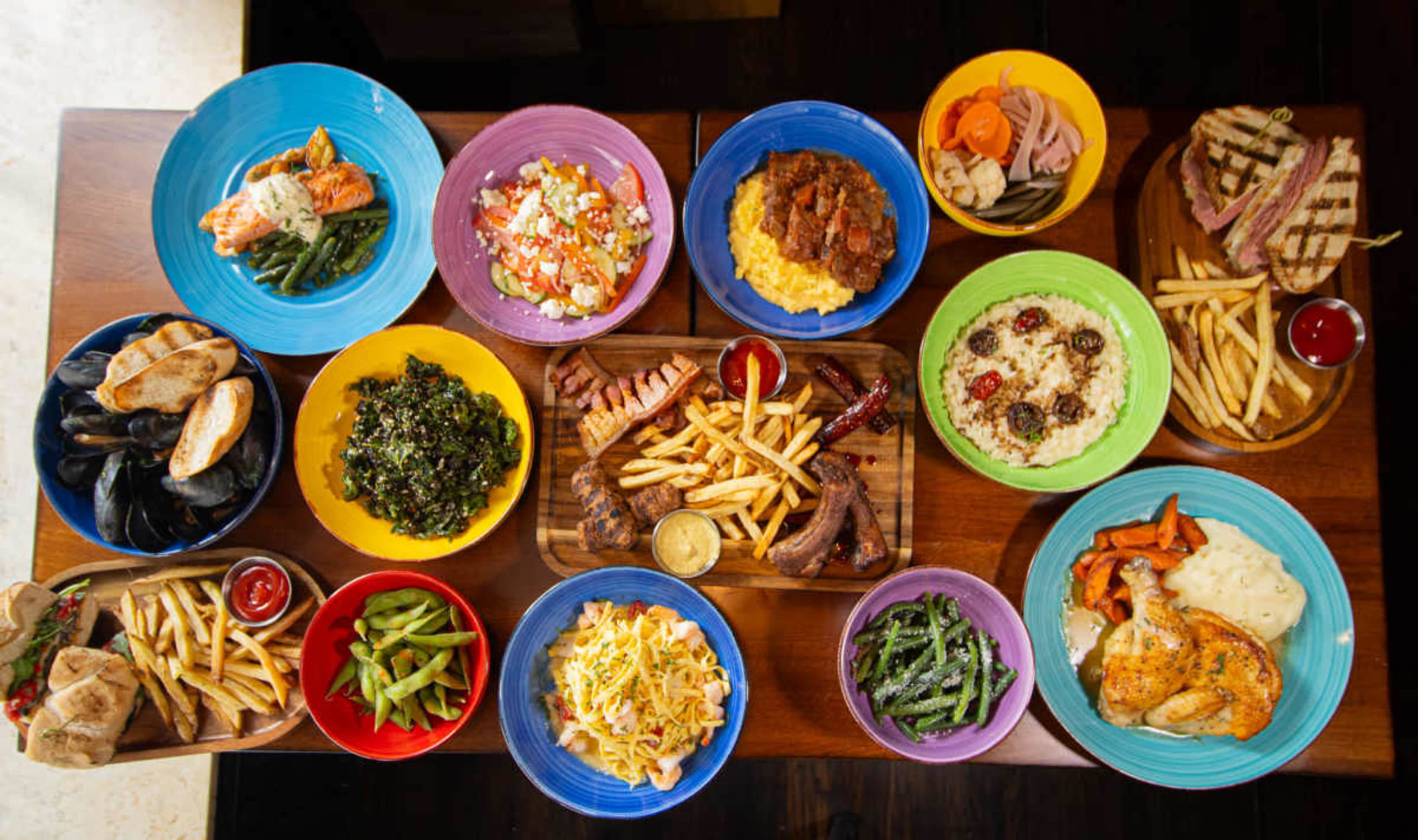 The image displays a variety of colorful plates arranged on a table, featuring different dishes including meats, vegetables, and sides.