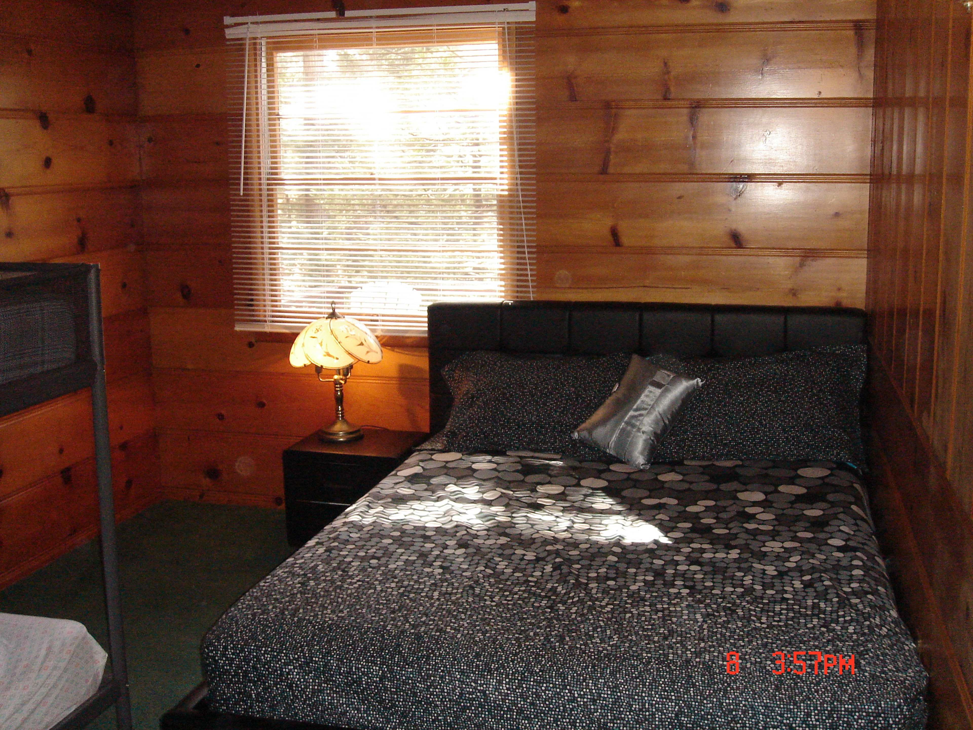 A bedroom features a bed with a patterned black and white comforter, a nightstand with a lamp, and a window with blinds allowing light to enter.