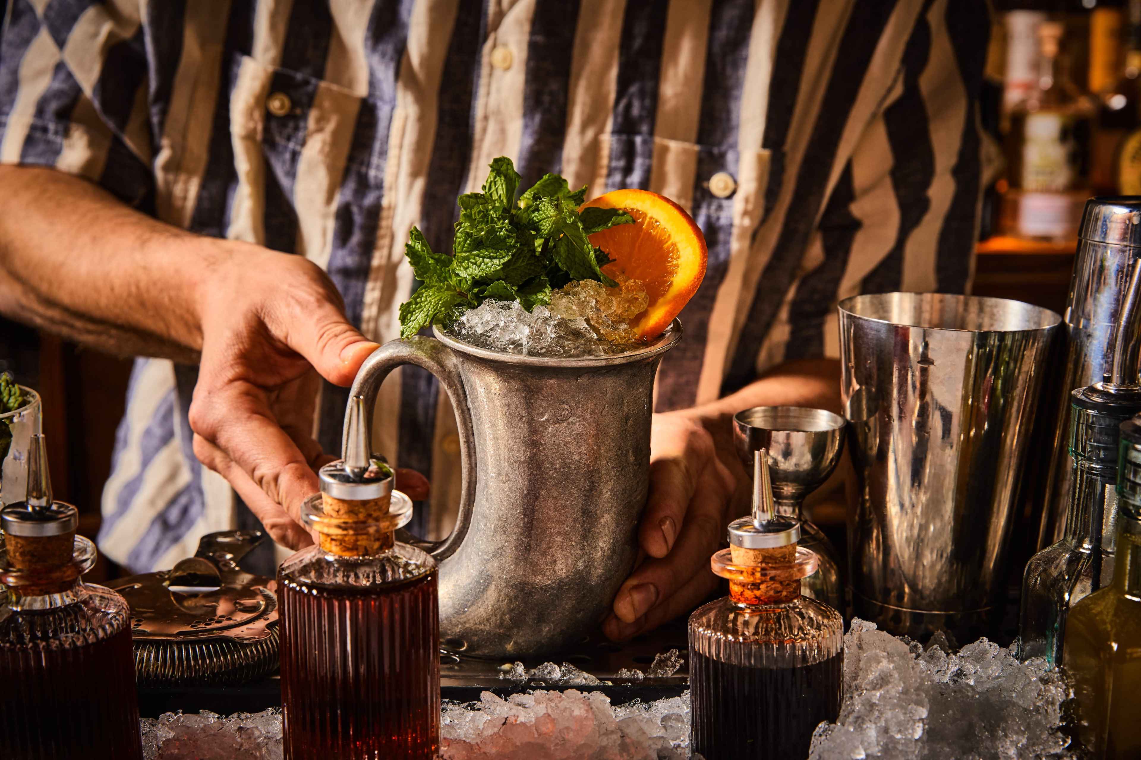 A bartender in a striped shirt holds a silver cup filled with ice, garnished with mint and an orange slice, amidst various bottles and tools on the bar.
