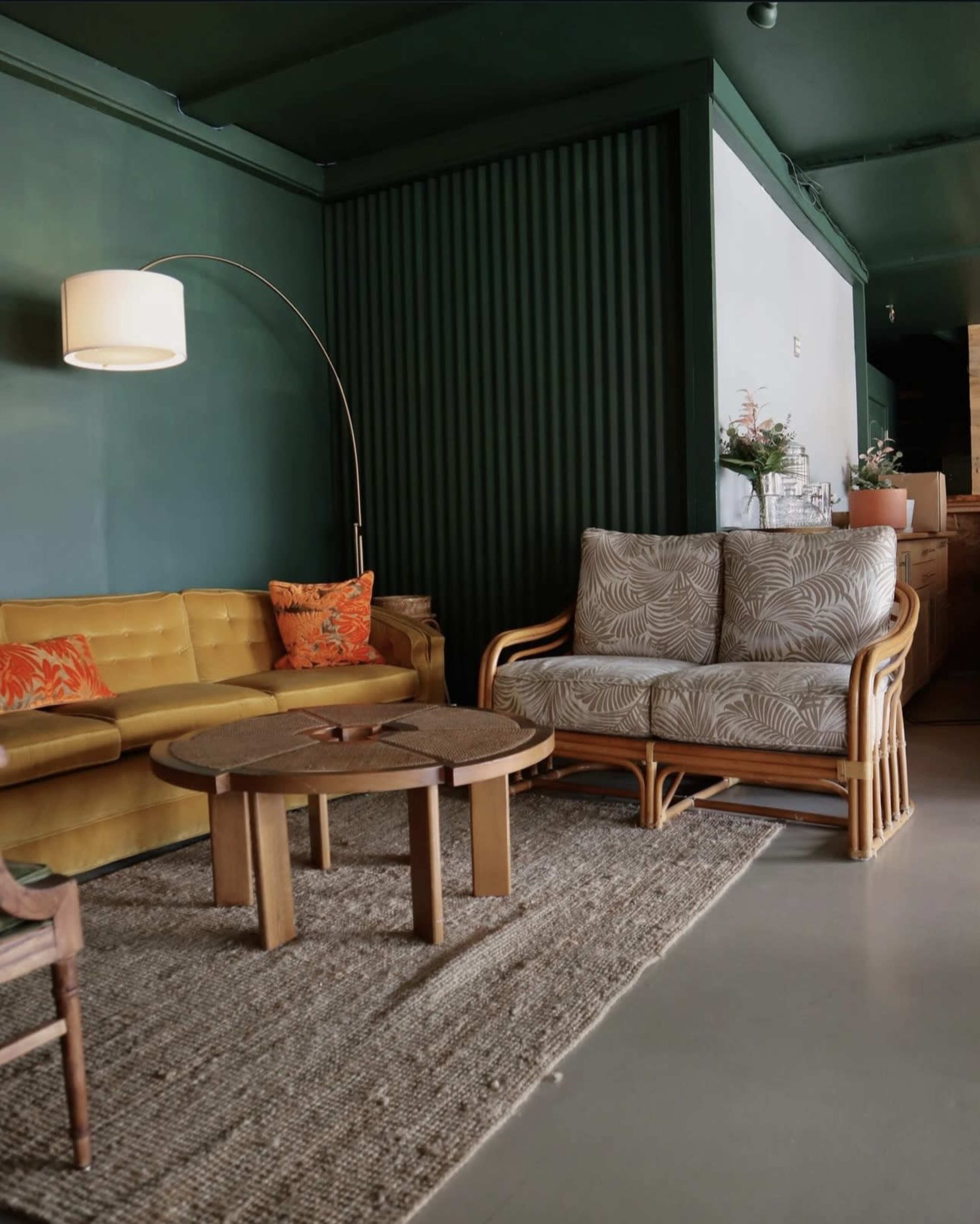 The image depicts a cozy lounge area featuring a yellow sofa, a patterned armchair, a round wooden coffee table, and a floor lamp against dark green walls.
