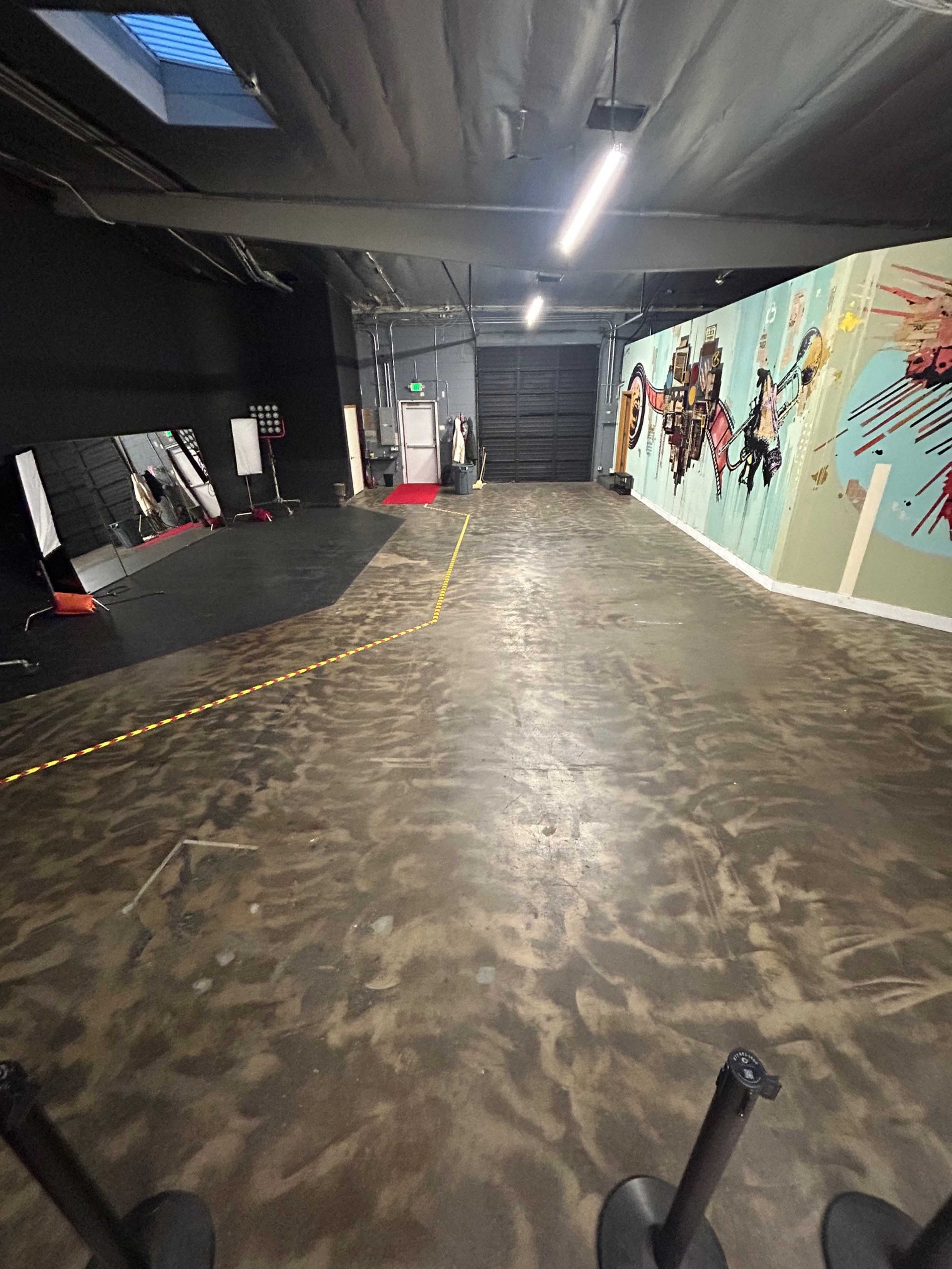 The image shows a spacious, dimly lit interior with a polished concrete floor, a large mural on one wall, and a mirrored area to the side.