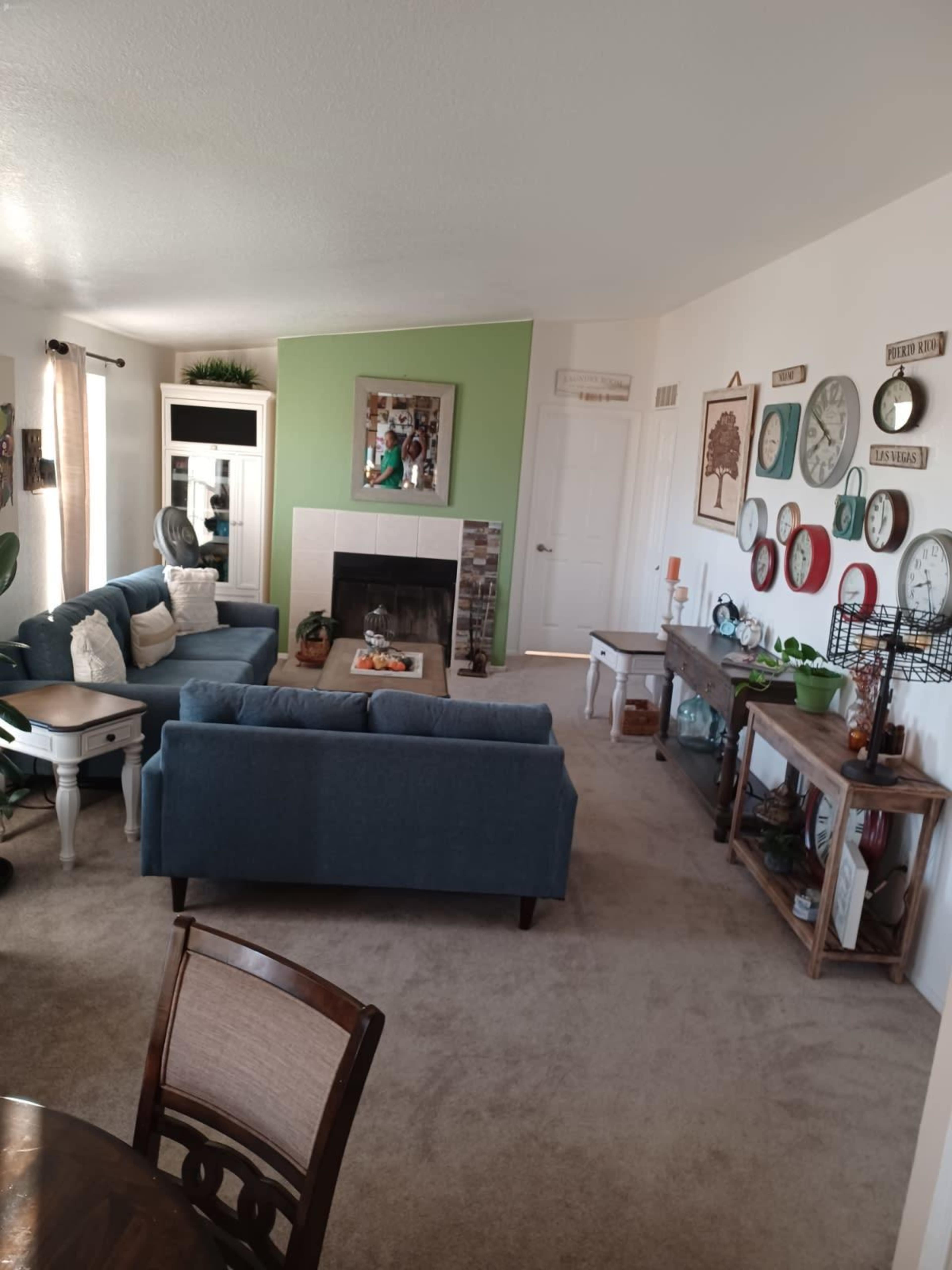 A living room features a fireplace, blue sofas, a decorative wall with various clocks, and wooden furniture.