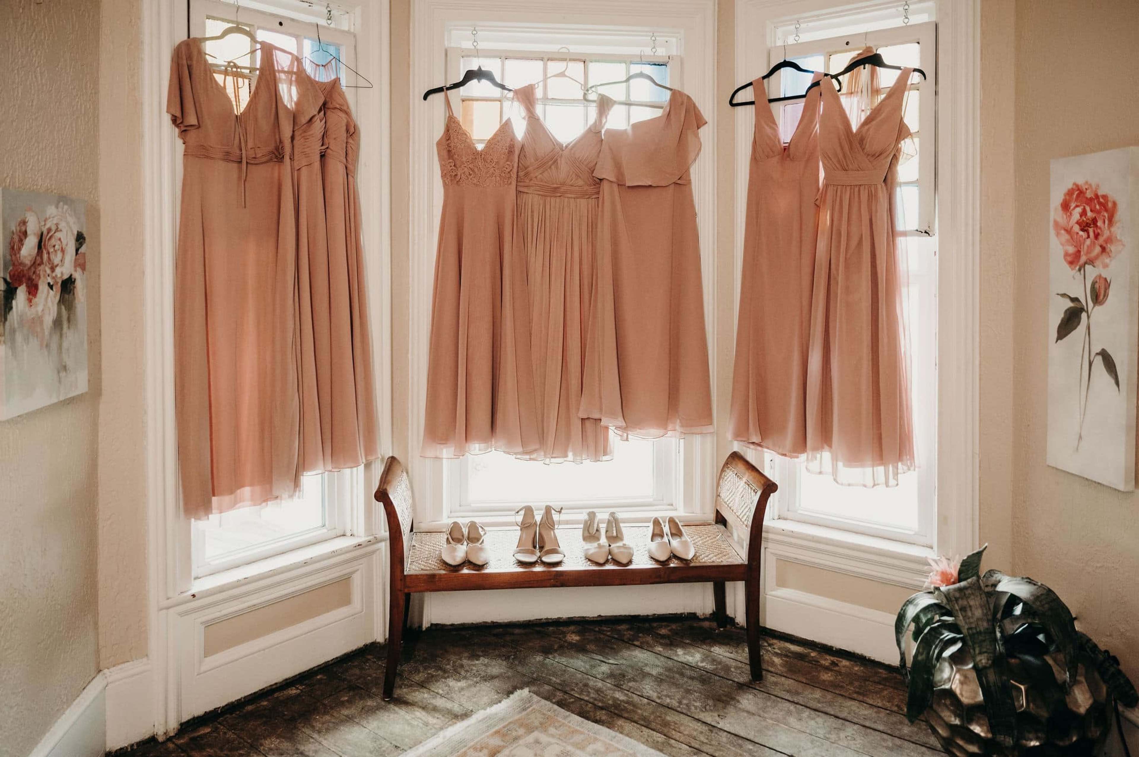 A row of light pink dresses hangs against a window, with a pair of shoes placed on a wooden bench below.