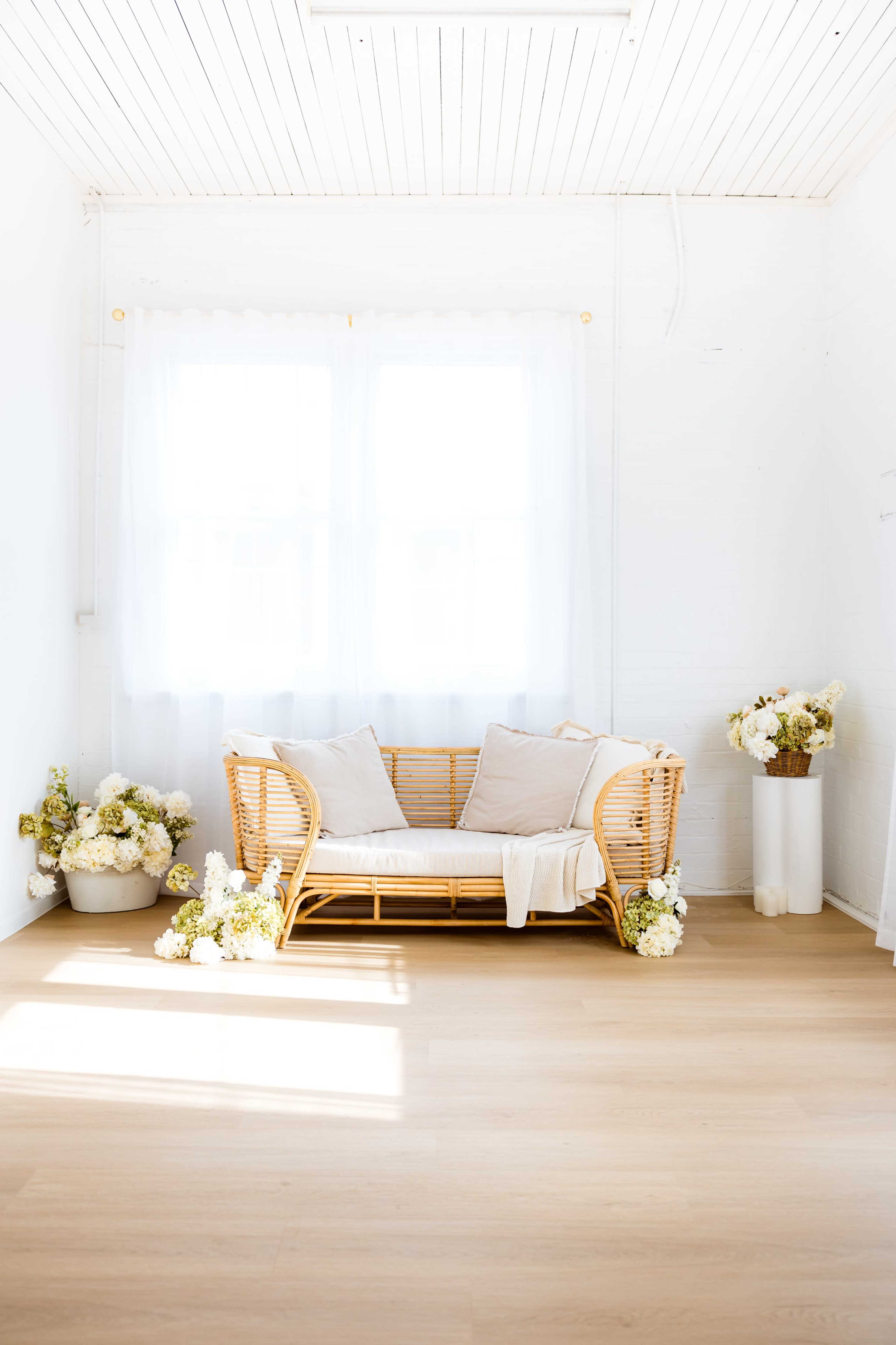 A rattan sofa with beige cushions is positioned in a bright room with white walls, complemented by potted flowers and ample natural light from a nearby window.