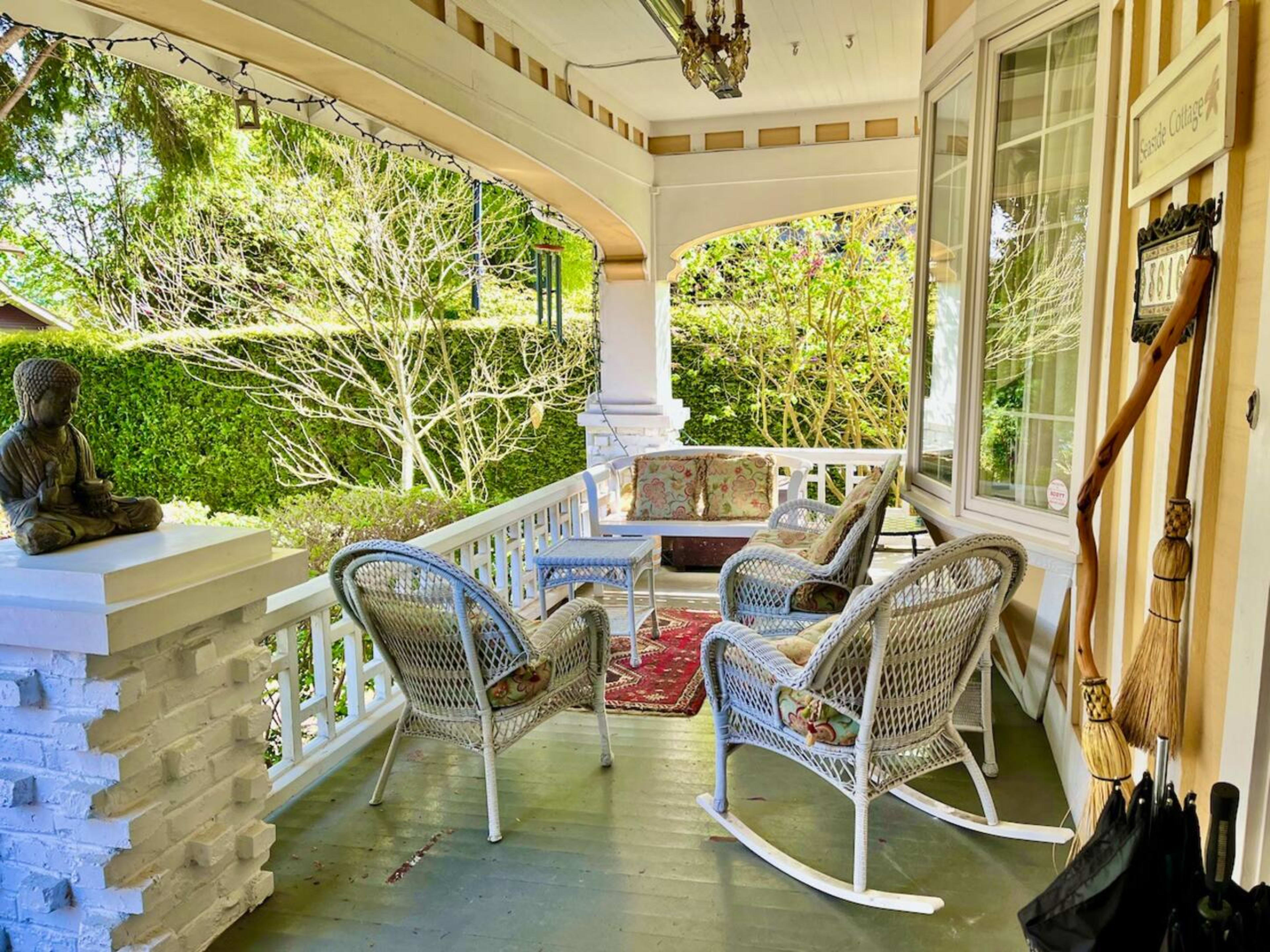 A porch with wicker chairs, a small table, and a decorative Buddha statue beside a couch, surrounded by greenery.
