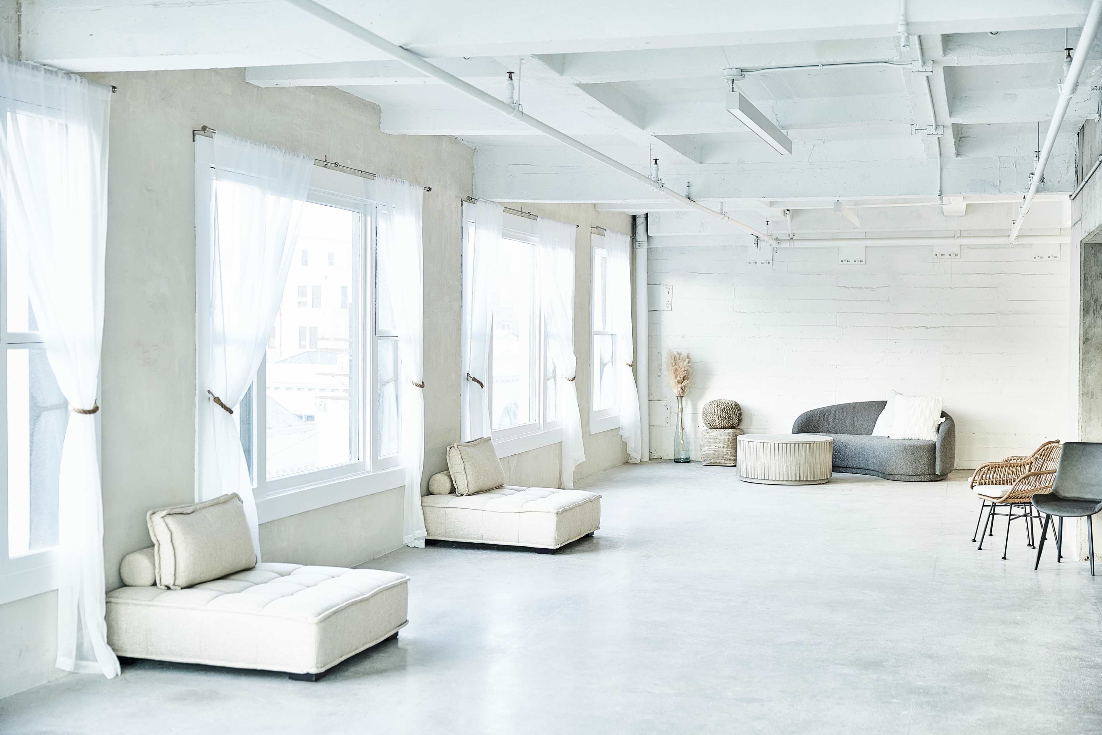 A bright, minimalist room features large windows, light-colored walls, and simple furniture arrangements, including two low sofas and a textured chair.
