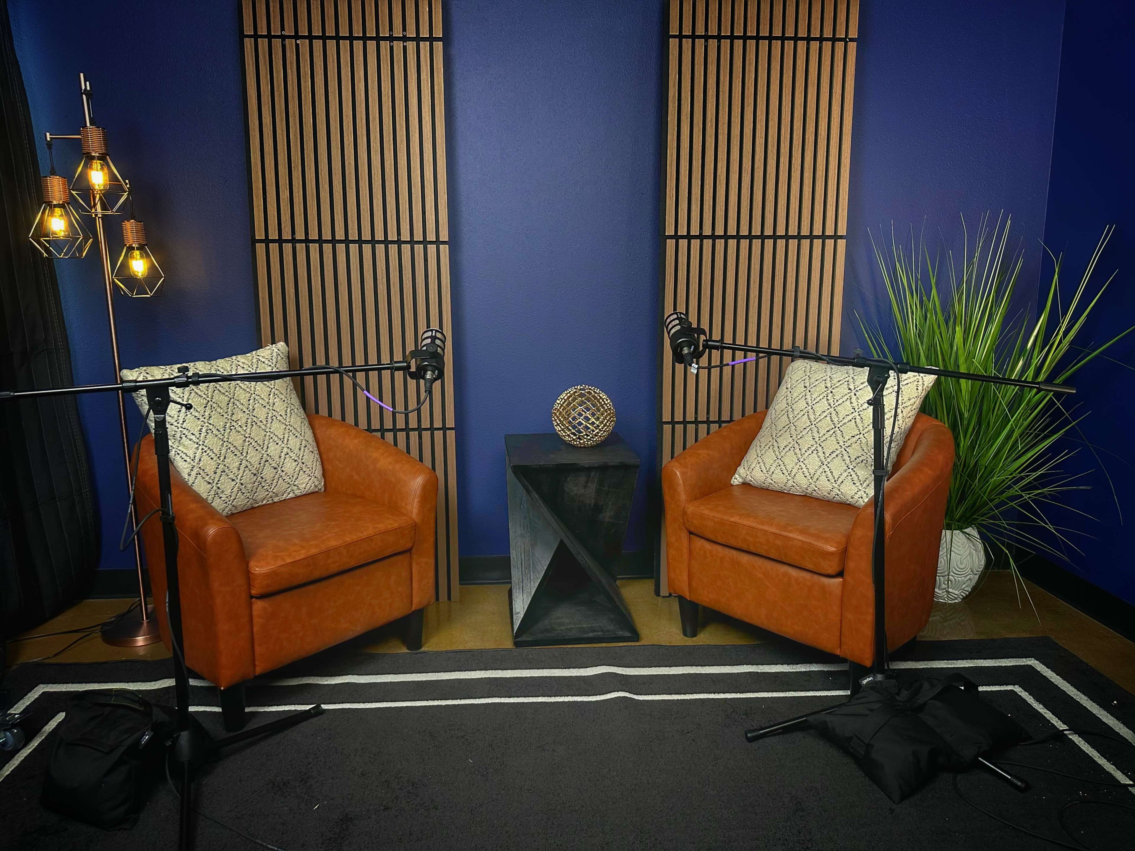 The image shows a cozy podcasting setup featuring two leather chairs, microphones mounted on stands, and decorative elements like a plant and a geometric object on a black pedestal table against a blue backdrop.