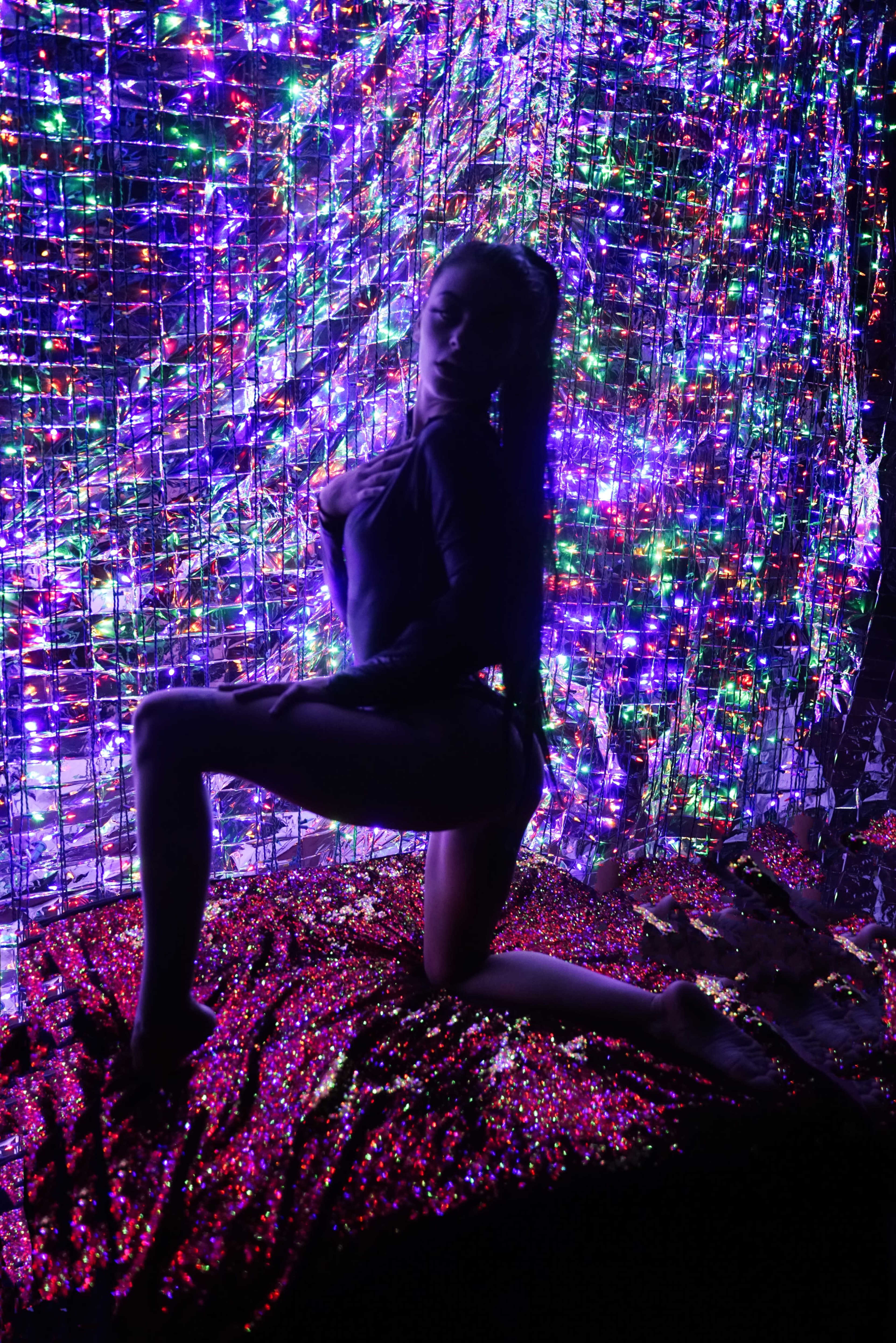 A person poses on a bed of sparkling material in front of a backdrop of colorful, illuminated lights.