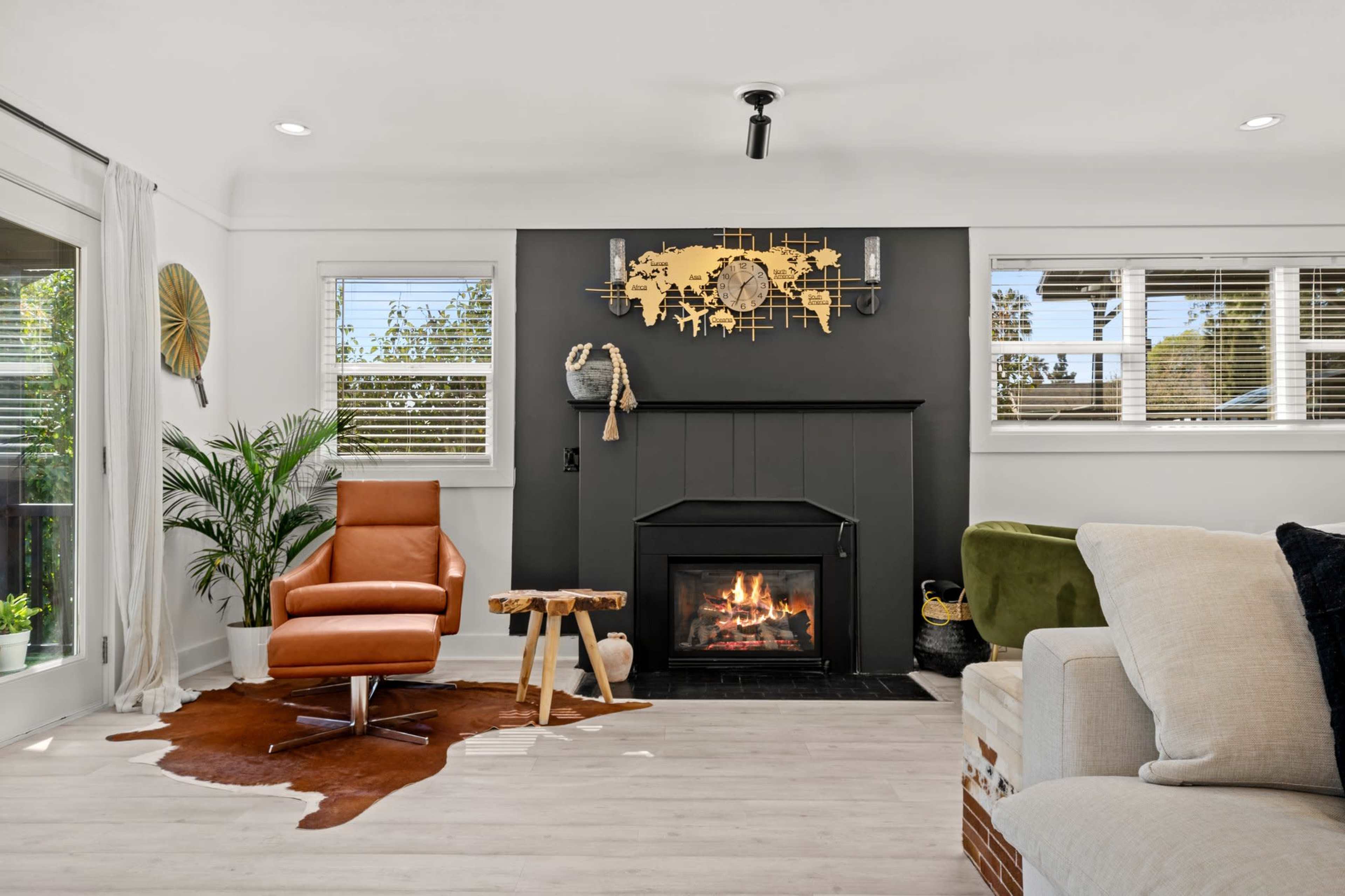 The living room features a modern design with a black fireplace, a large world map decoration above it, an orange leather chair, a cowhide rug, and natural light from multiple windows.