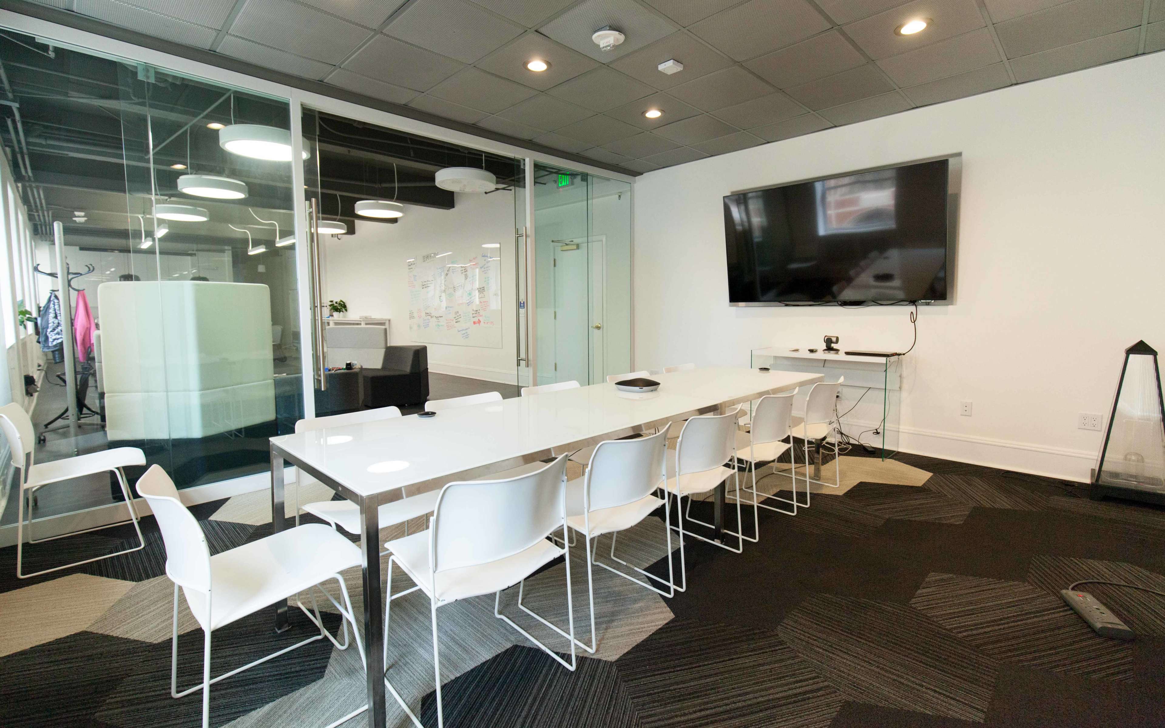 A modern conference room features a long white table with ten white chairs, a large wall-mounted screen, and glass walls displaying a collaborative space.
