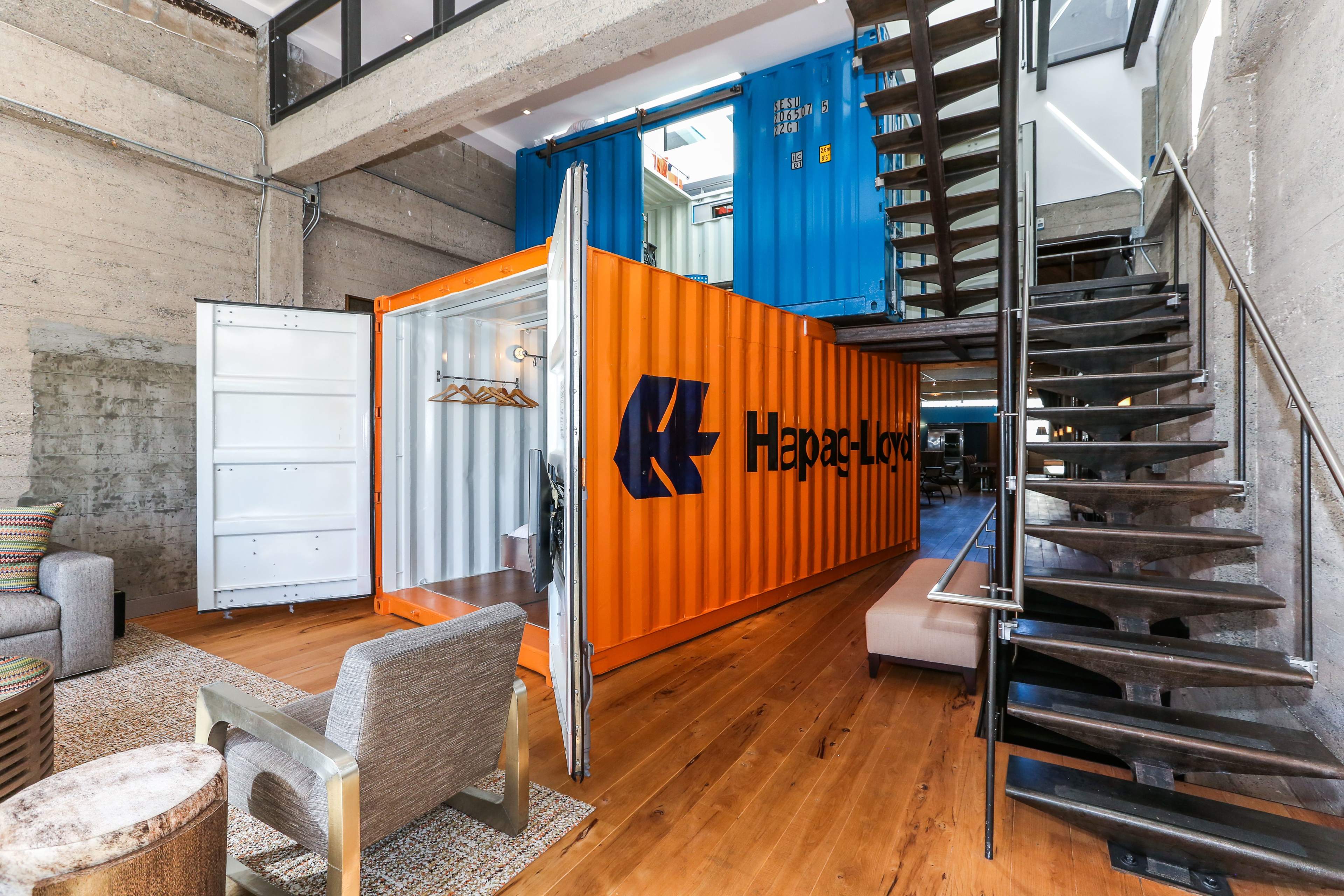 A modern interior space featuring two stacked shipping containers, one orange and one blue, with a staircase leading to an upper level.