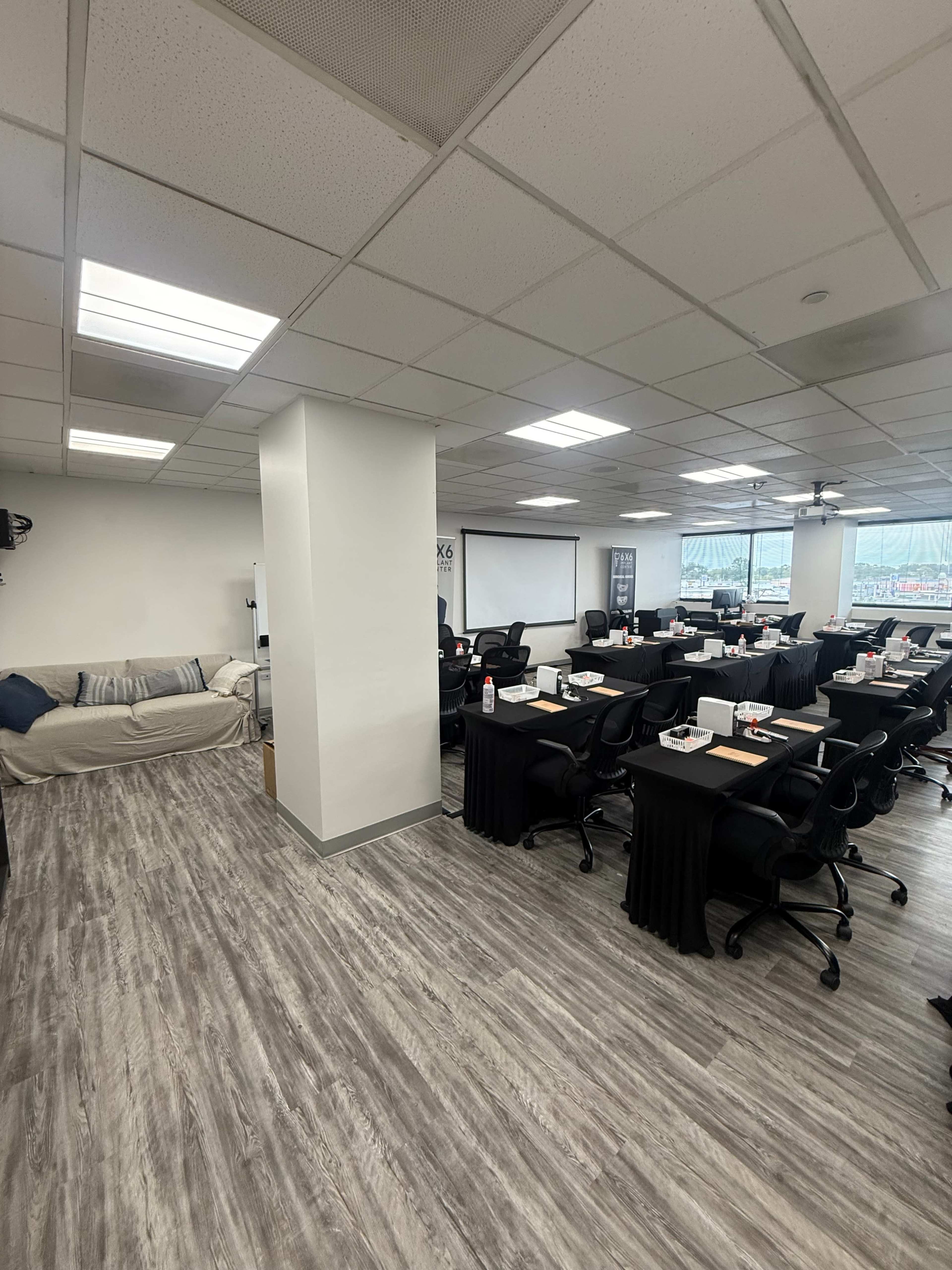 The image shows a modern classroom setup with several tables arranged for an instructional session, featuring black tablecloths, chairs, and a large window providing natural light.