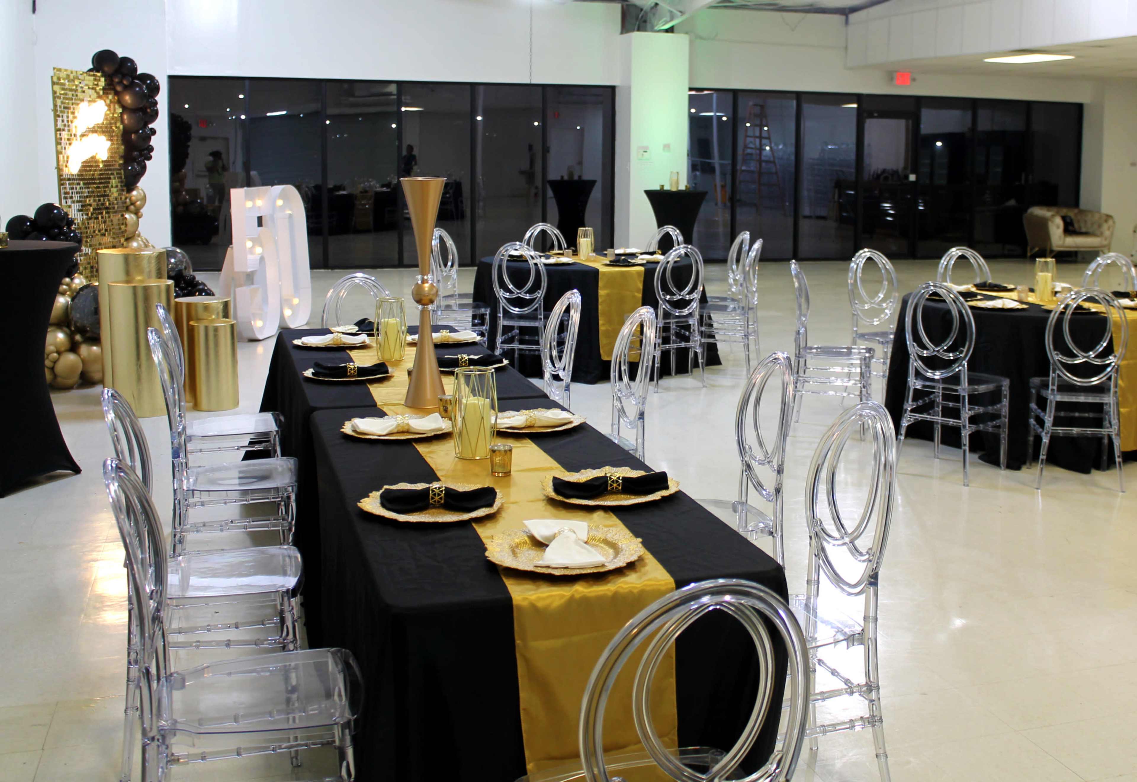 The image shows a decorated event space with tables set in black and gold, featuring clear plastic chairs and elegant tableware.