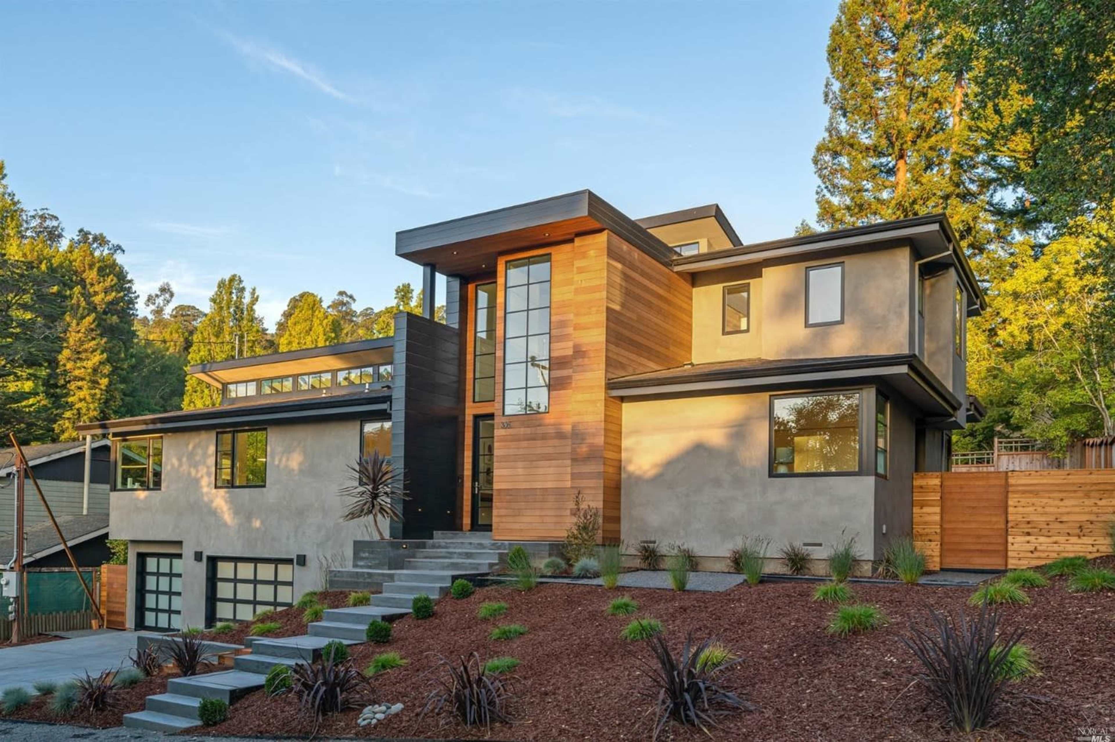 A modern two-story house with a wooden and concrete facade, surrounded by landscaped greenery and a stone pathway.