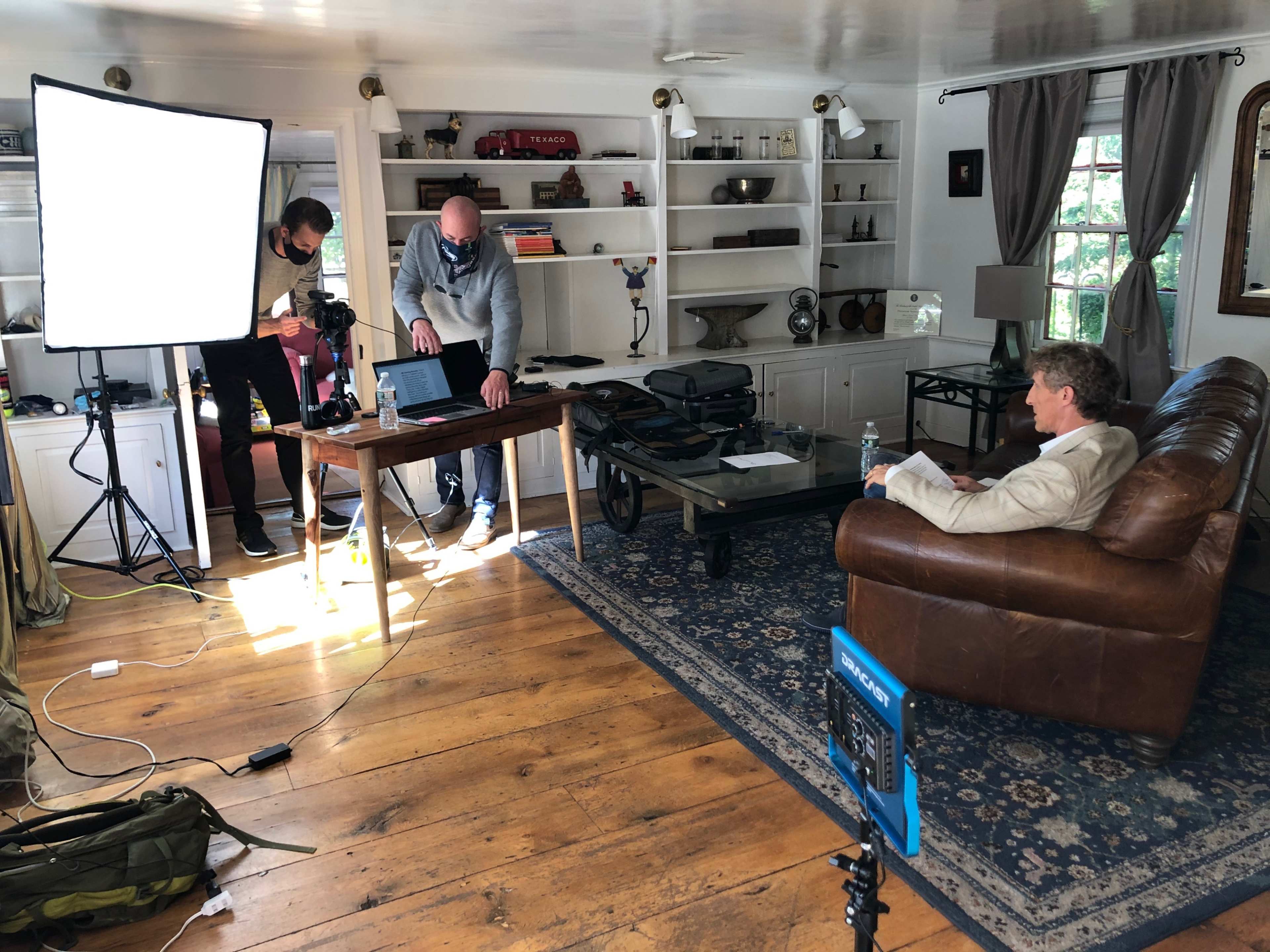 Two individuals are setting up equipment in a living room while a third person sits on a couch.